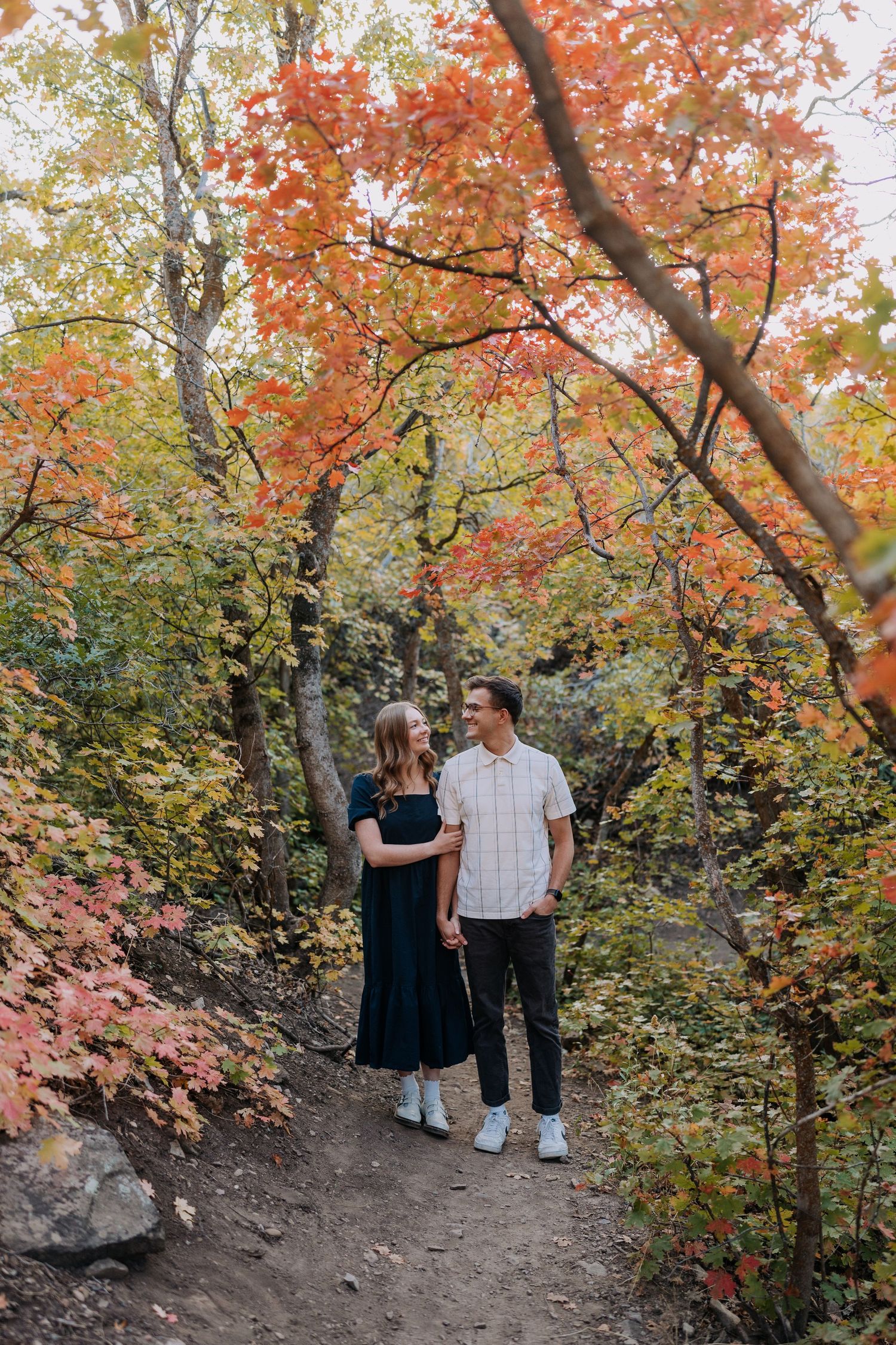 Couple holding hands on a forest trail surrounded by vibrant red and orange autumn foliage.