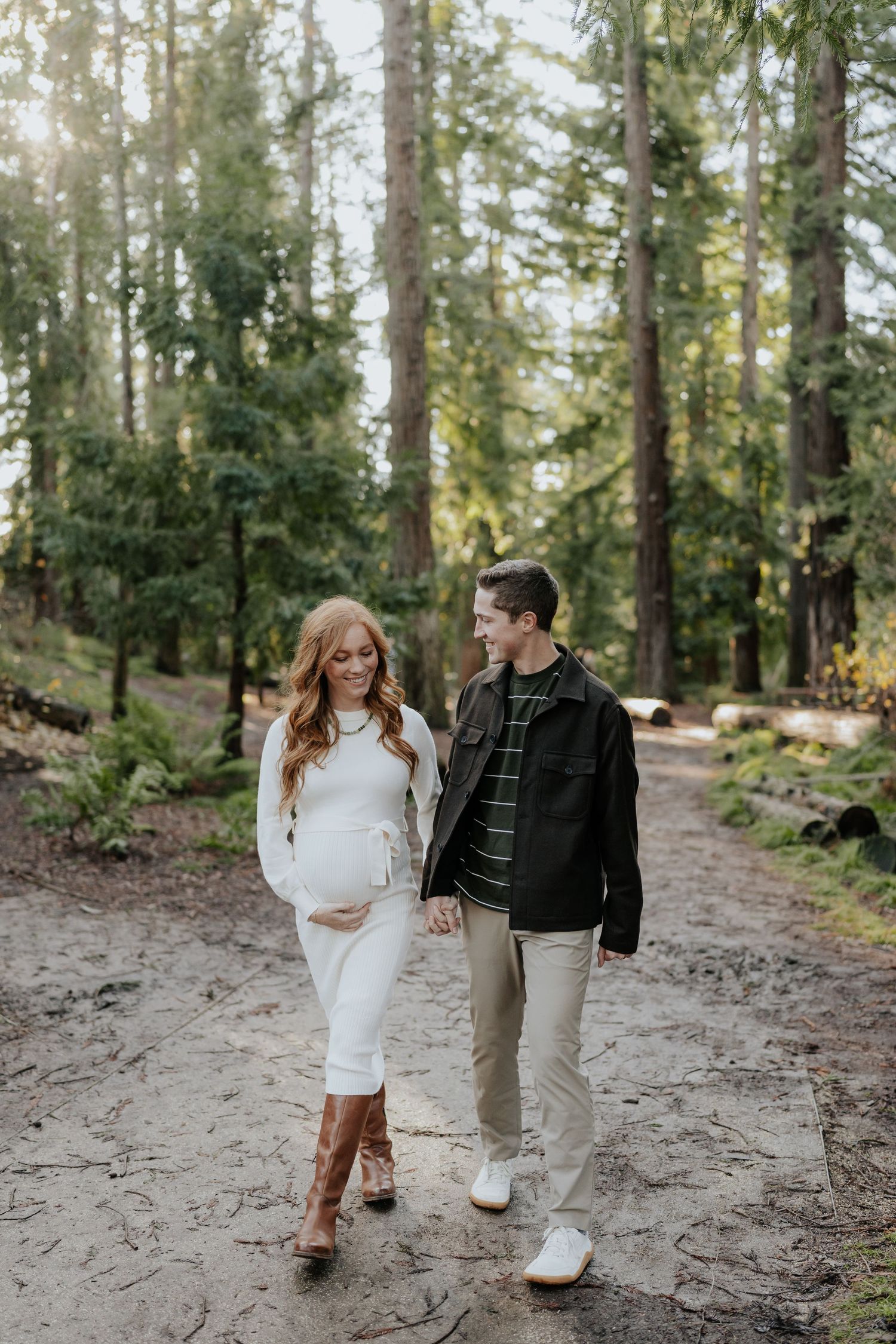 Pregnant couple walking hand-in-hand on a sunlit forest path, woman in white dress and boots in Davis, California.