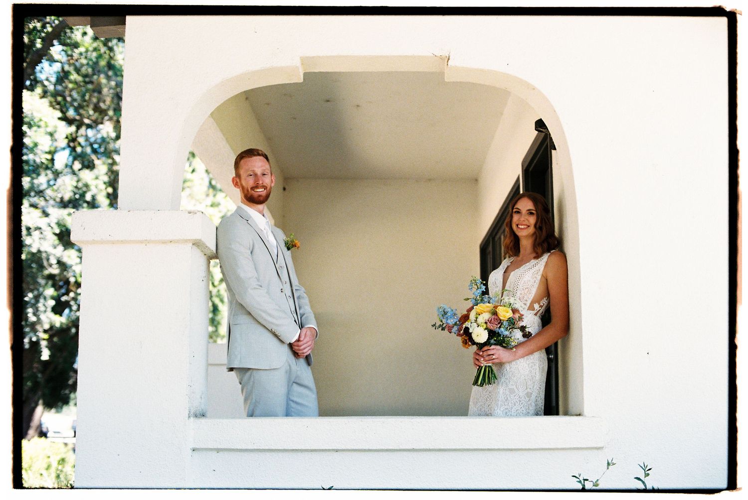 Bride holding yellow and blue bouquet stands in doorway while groom in gray suit leans against white arch outdoors.