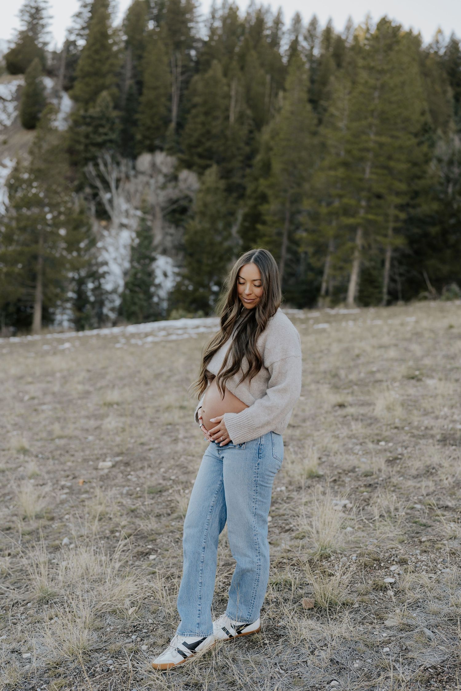 Pregnant woman in beige cropped sweater and jeans cradling baby bump in mountain meadow.
