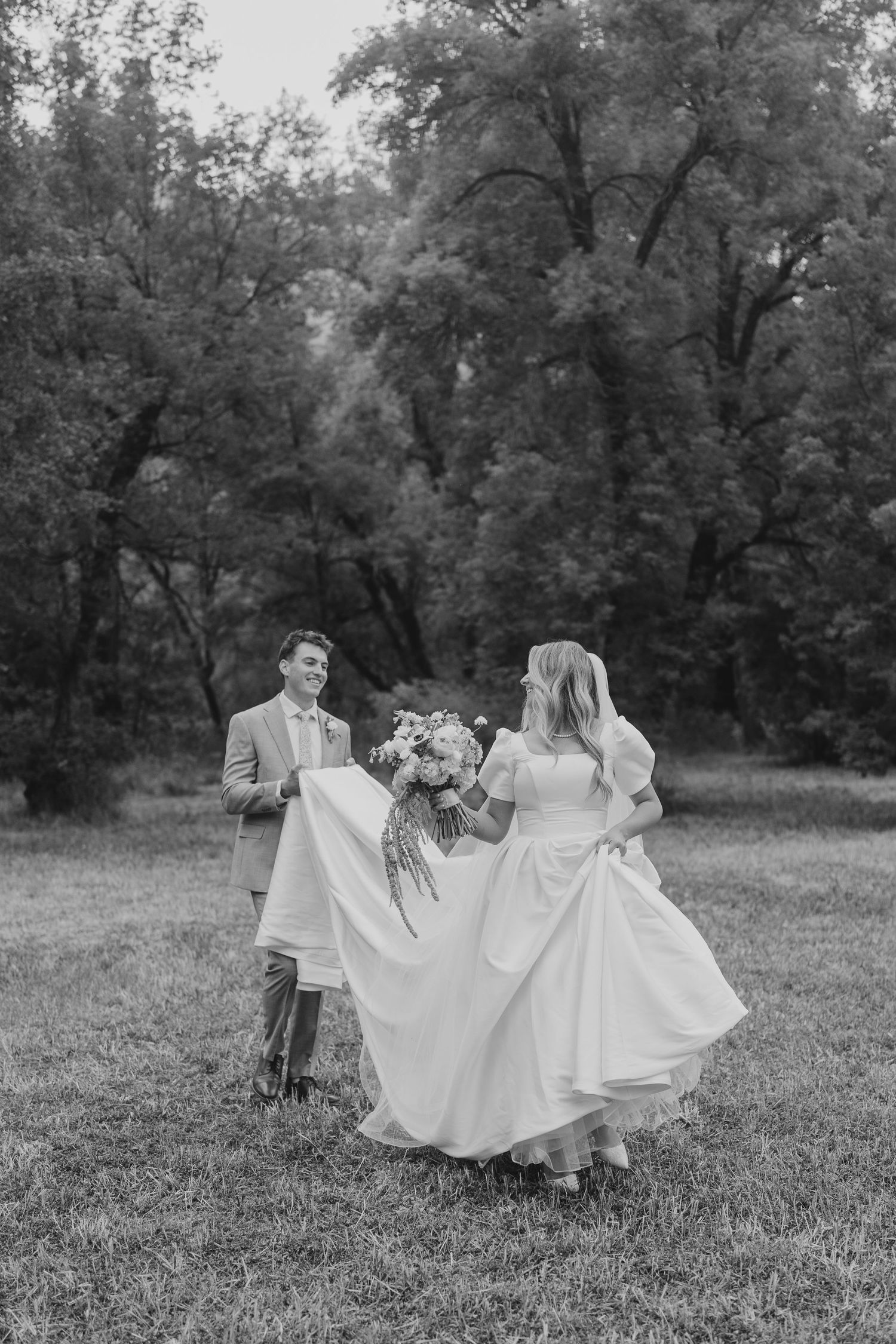 Black and white photo of a smiling groom lifting his bride's dress as she walks through a meadow near Provo, Utah.