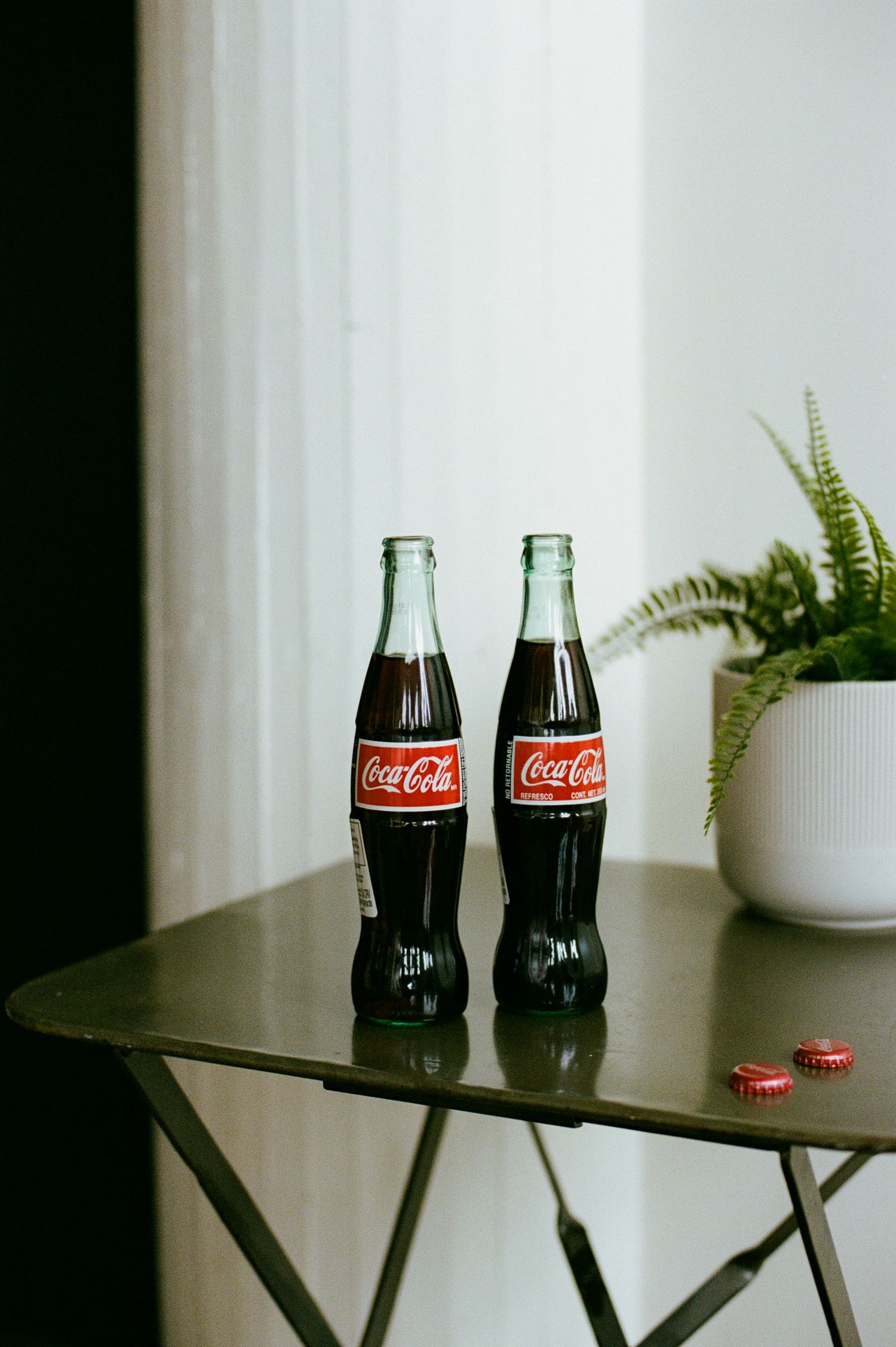 Two classic Coca-Cola glass bottles on a metal table beside a fern plant near a window.