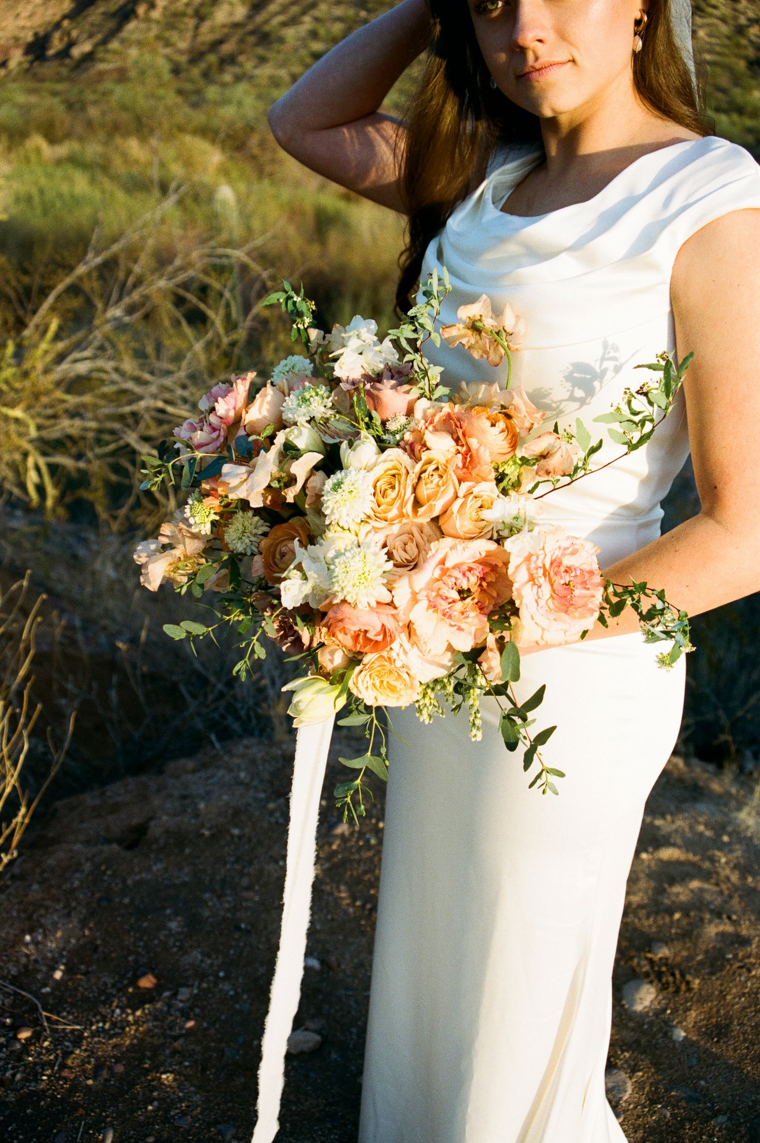 Bride in white gown holds lush peach and blush cascading bouquet in desert setting.