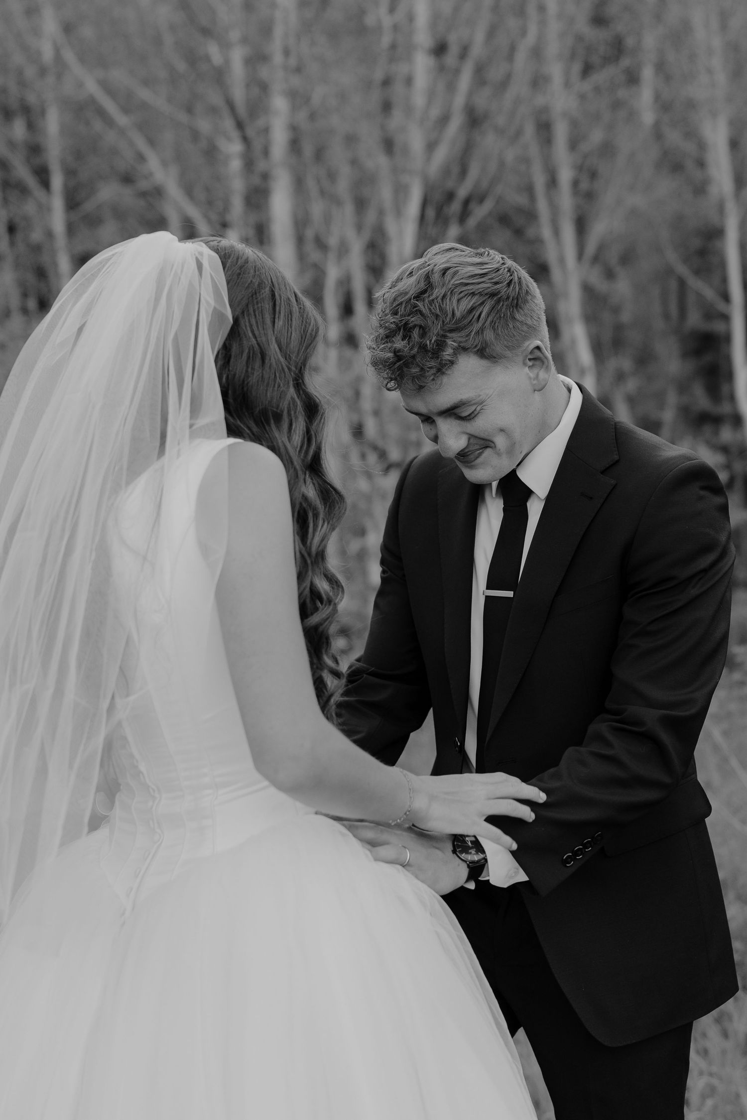 Black and white photo of a bride and groom holding hands outdoors, groom in suit smiling down at bride in wedding dress and veil.