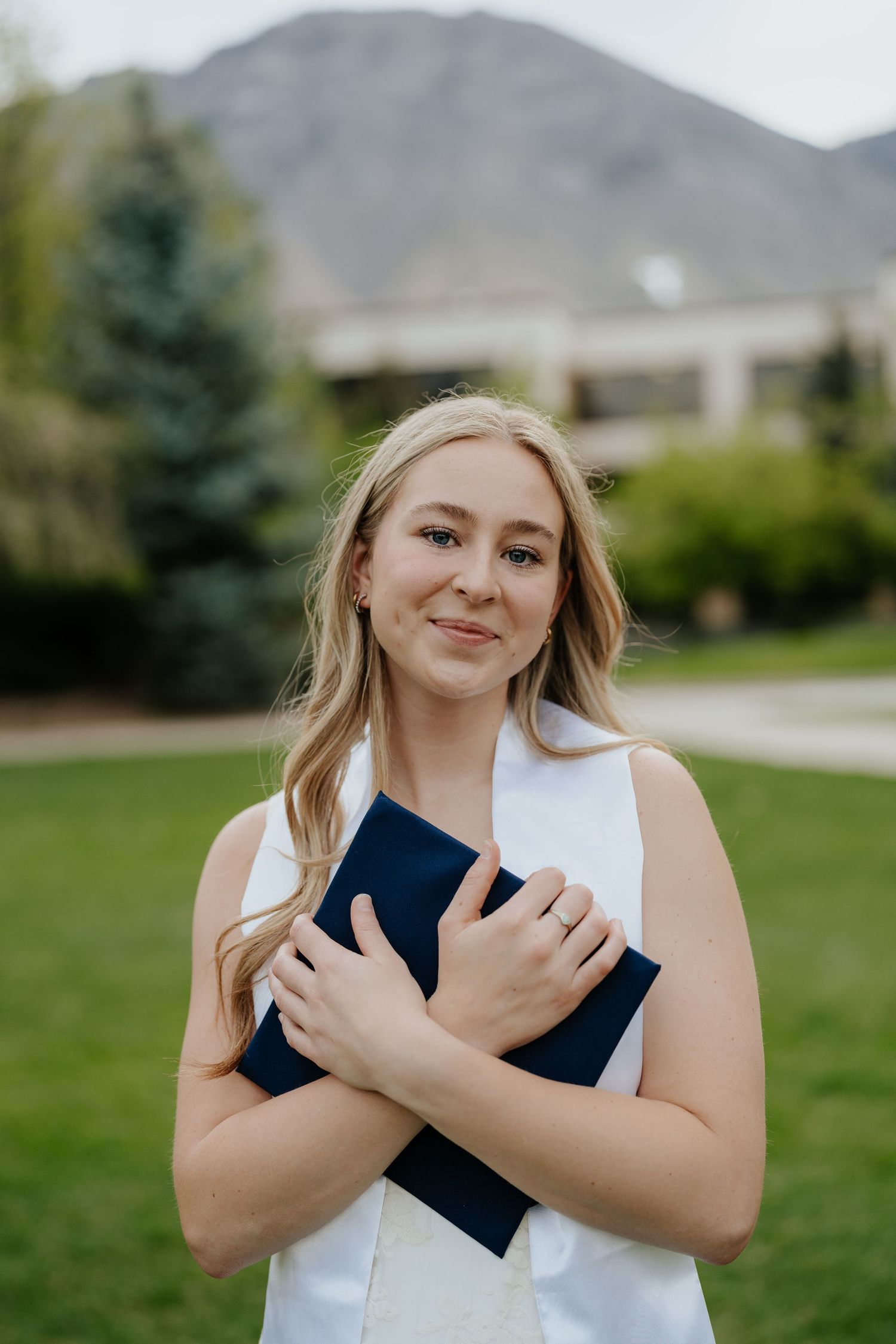 Blonde BYU graduate in white dress hugging navy cap outdoors with mountain backdrop.