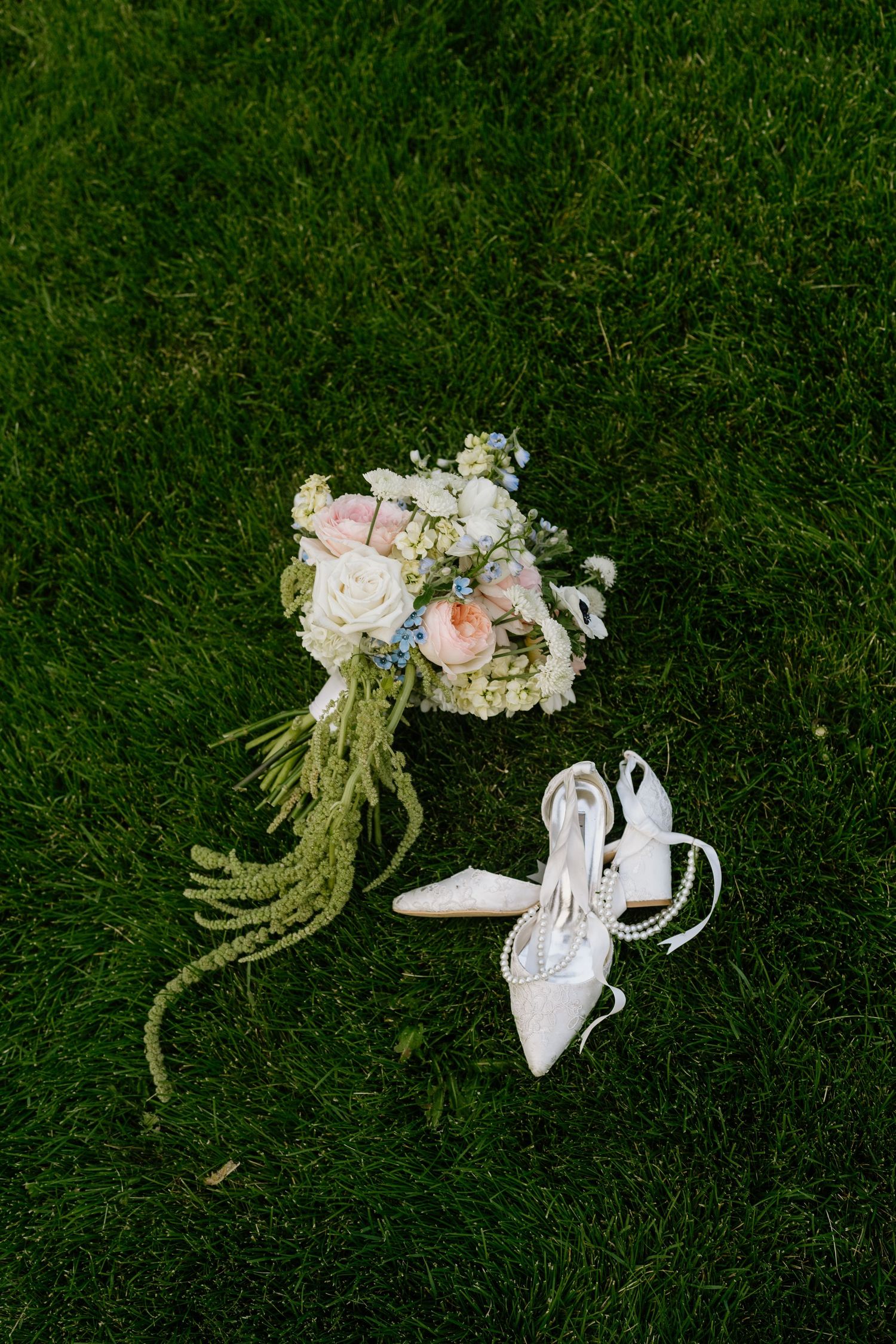 Elegant bridal bouquet with pink and white flowers beside pearl-adorned white heels on green grass.