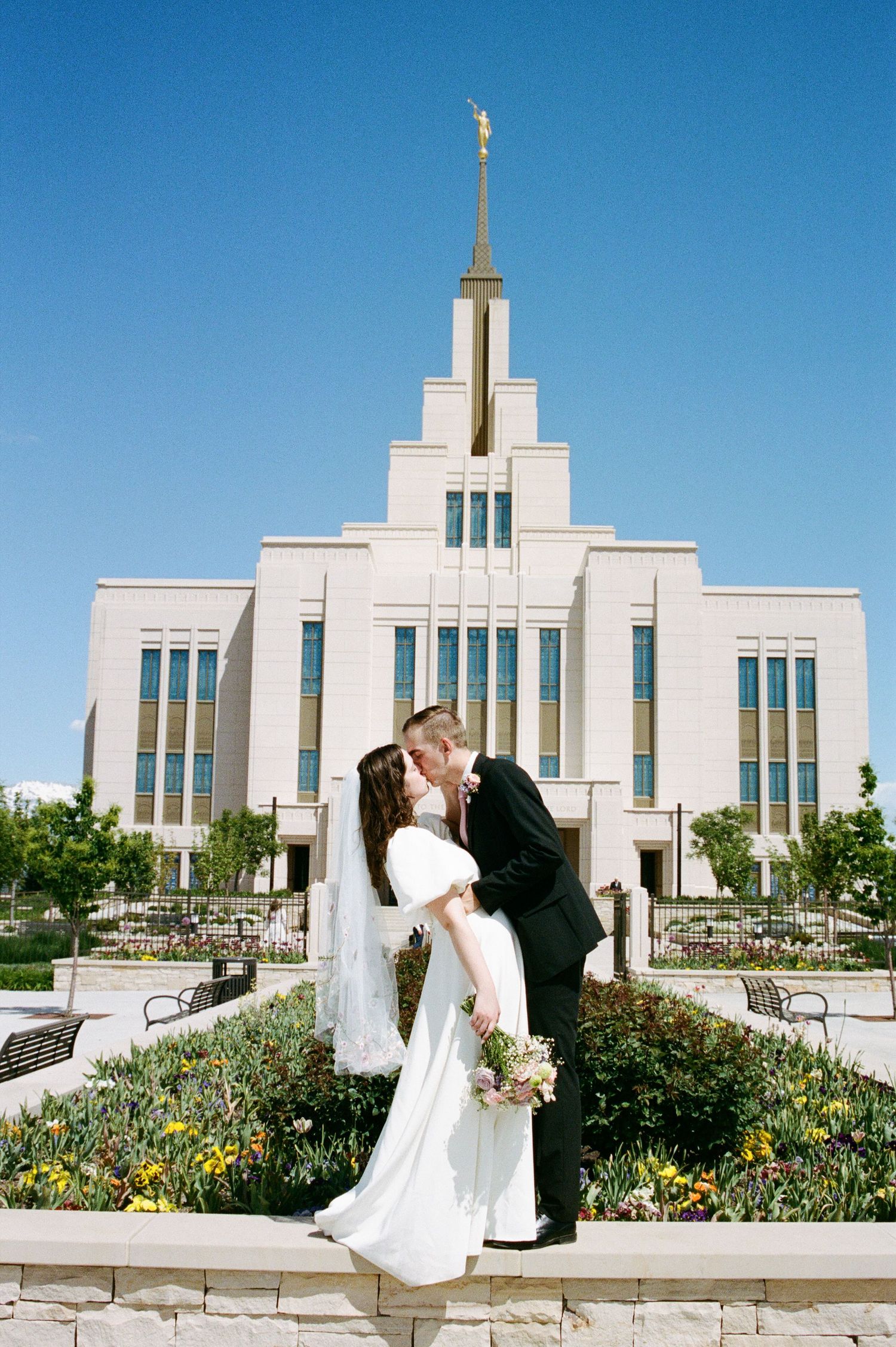 Bride and groom kissing in front of a white LDS temple under a bright blue sky.