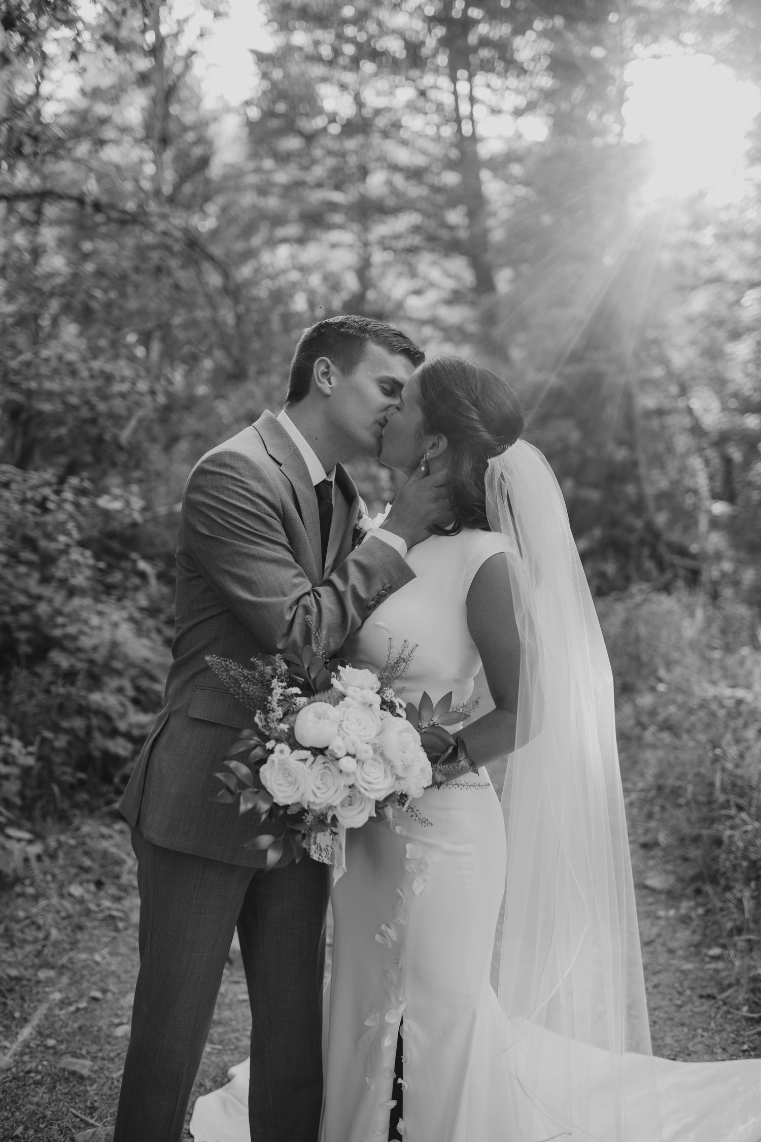 Black and white romantic kiss between bride and groom in sunlit forest with bouquet near Provo, Utah.