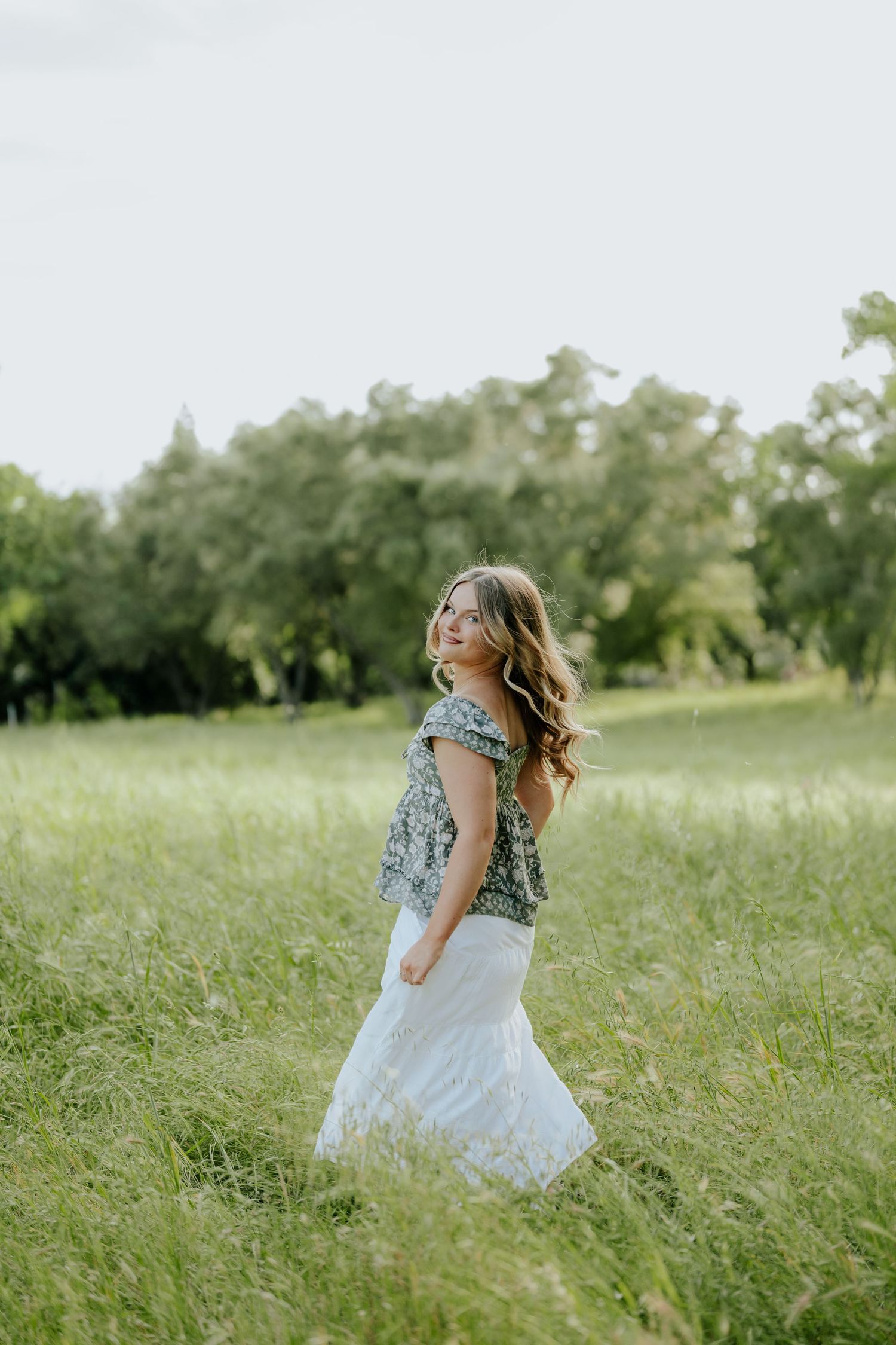 Young woman in floral top and white skirt walking through lush green meadow during golden hour near Sacramento, California.