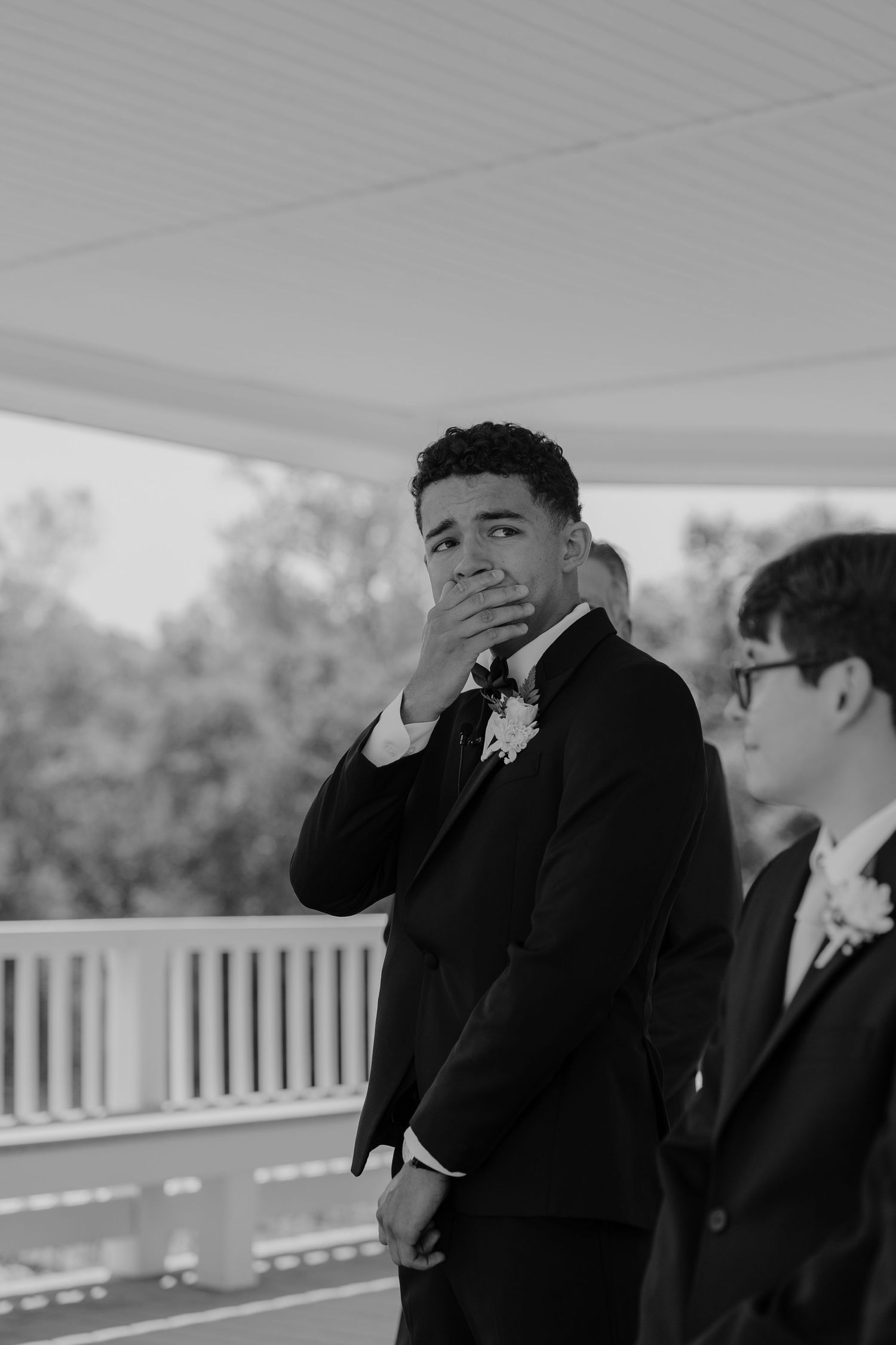 Black and white photo of emotional groom covering mouth at wedding altar, wearing suit with boutonniere.