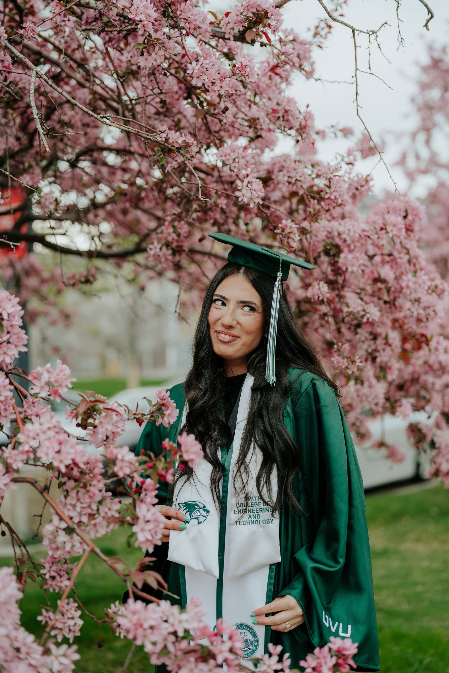 Young woman in green graduation cap and gown posing among pink cherry blossoms in spring.