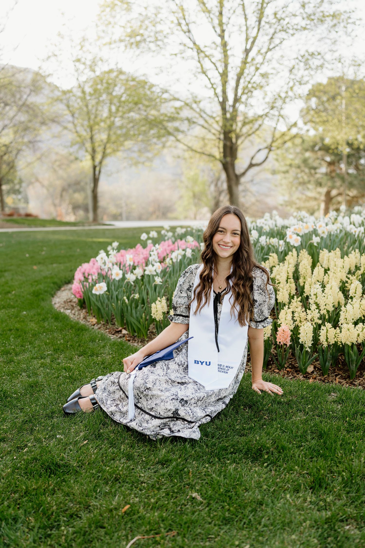 BYU graduate in floral dress with white stole sitting on grass beside colorful spring flowers.