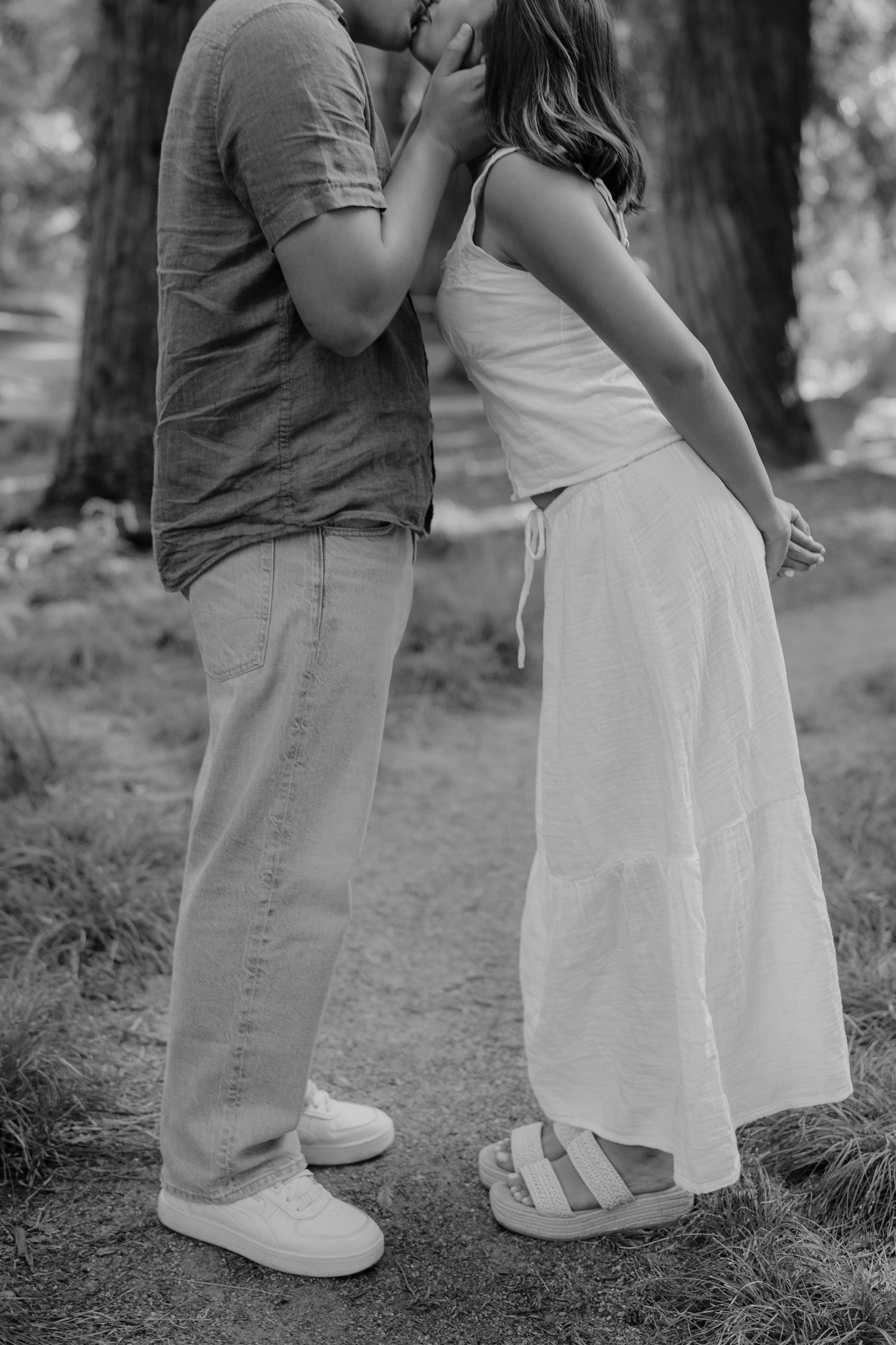 Black and white photo of a couple sharing a kiss outdoors, woman in white dress and sandals, man in casual shirt and jeans.