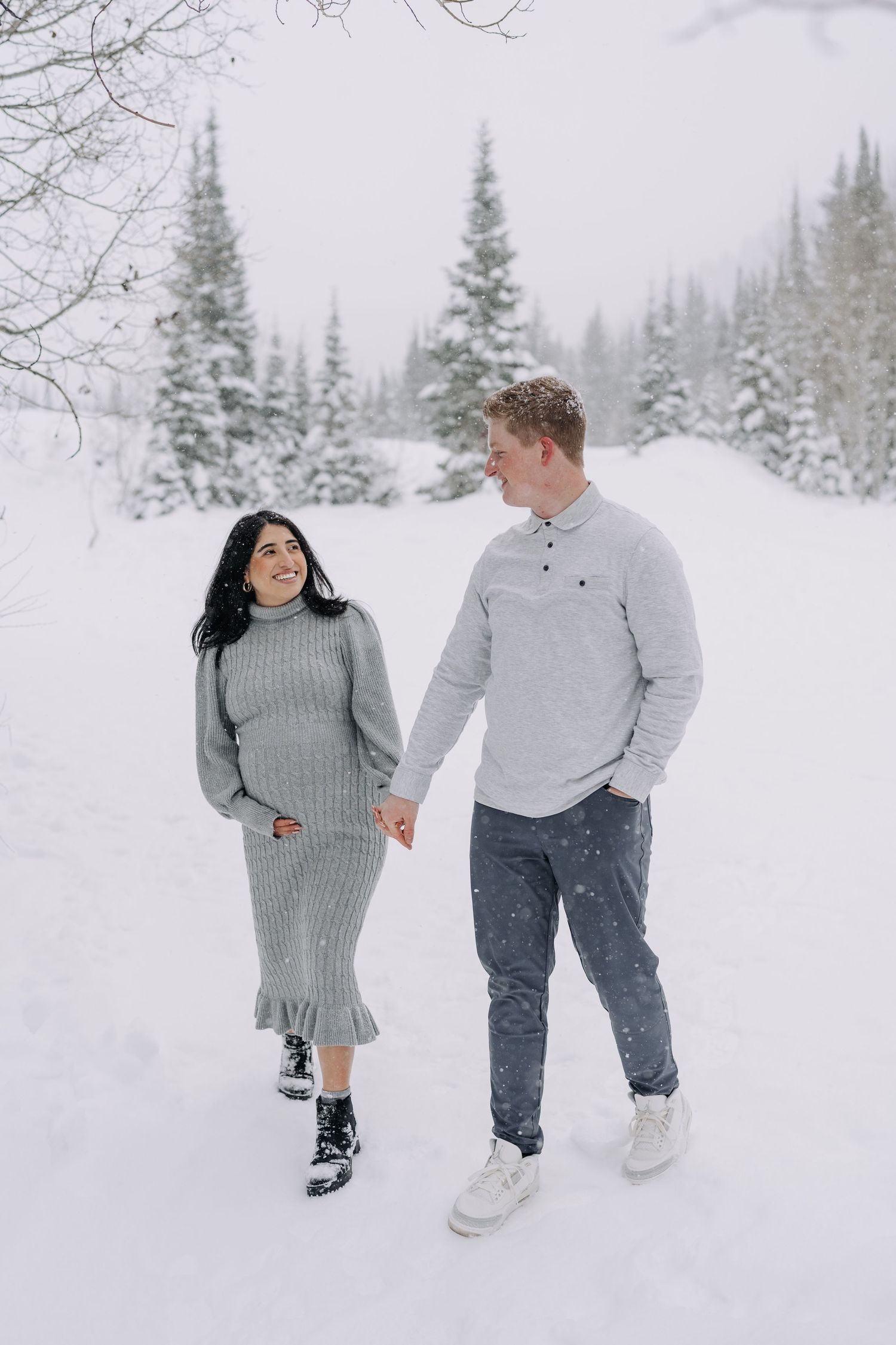 Pregnant couple holding hands walking through snowy winter forest, wearing matching gray outfits.