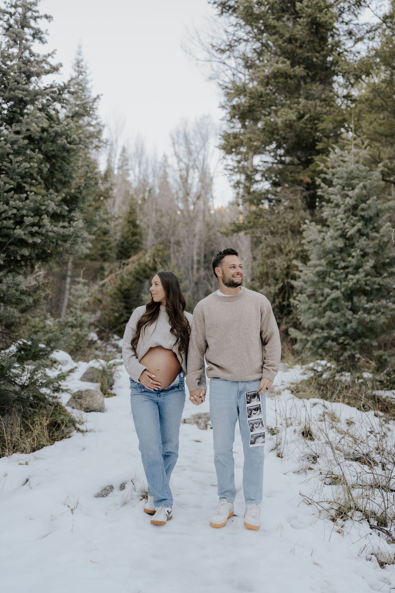 Expecting couple walking hand-in-hand on snowy forest path, man holding ultrasound photos.