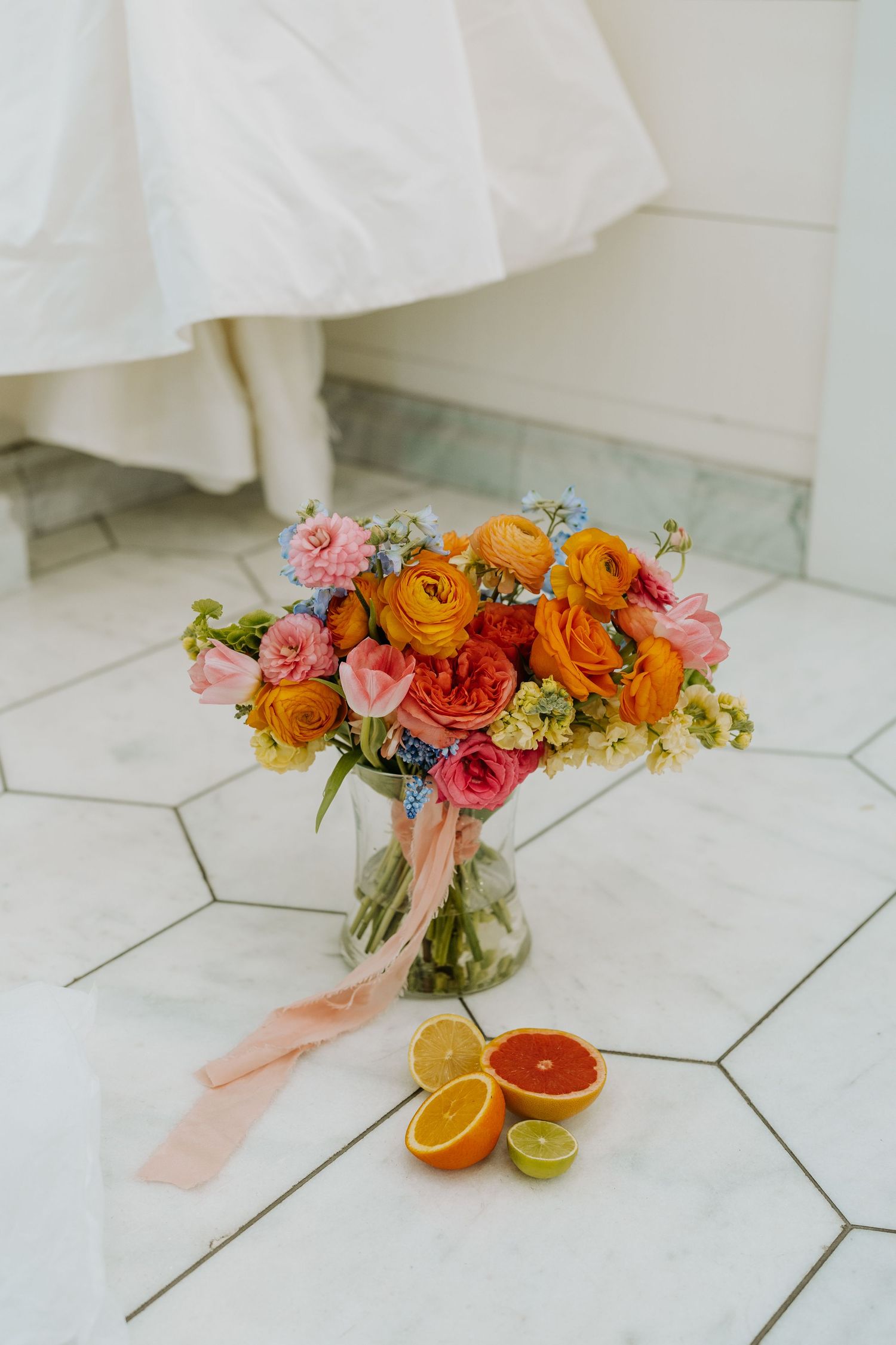 Colorful bridal bouquet with orange and pink ranunculus in glass vase beside sliced citrus on marble tile floor.