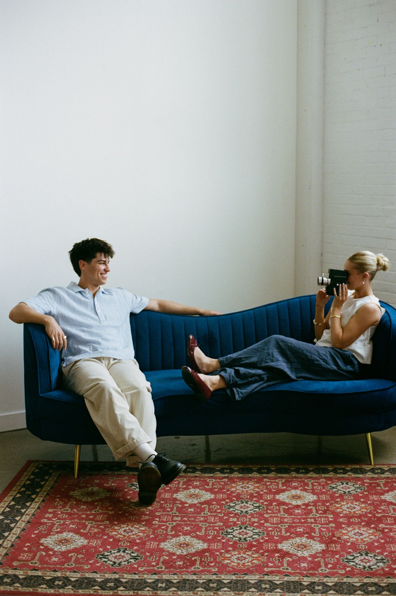 A man and woman relax on a blue velvet sofa on a red rug, as she photographs him with a vintage camera.