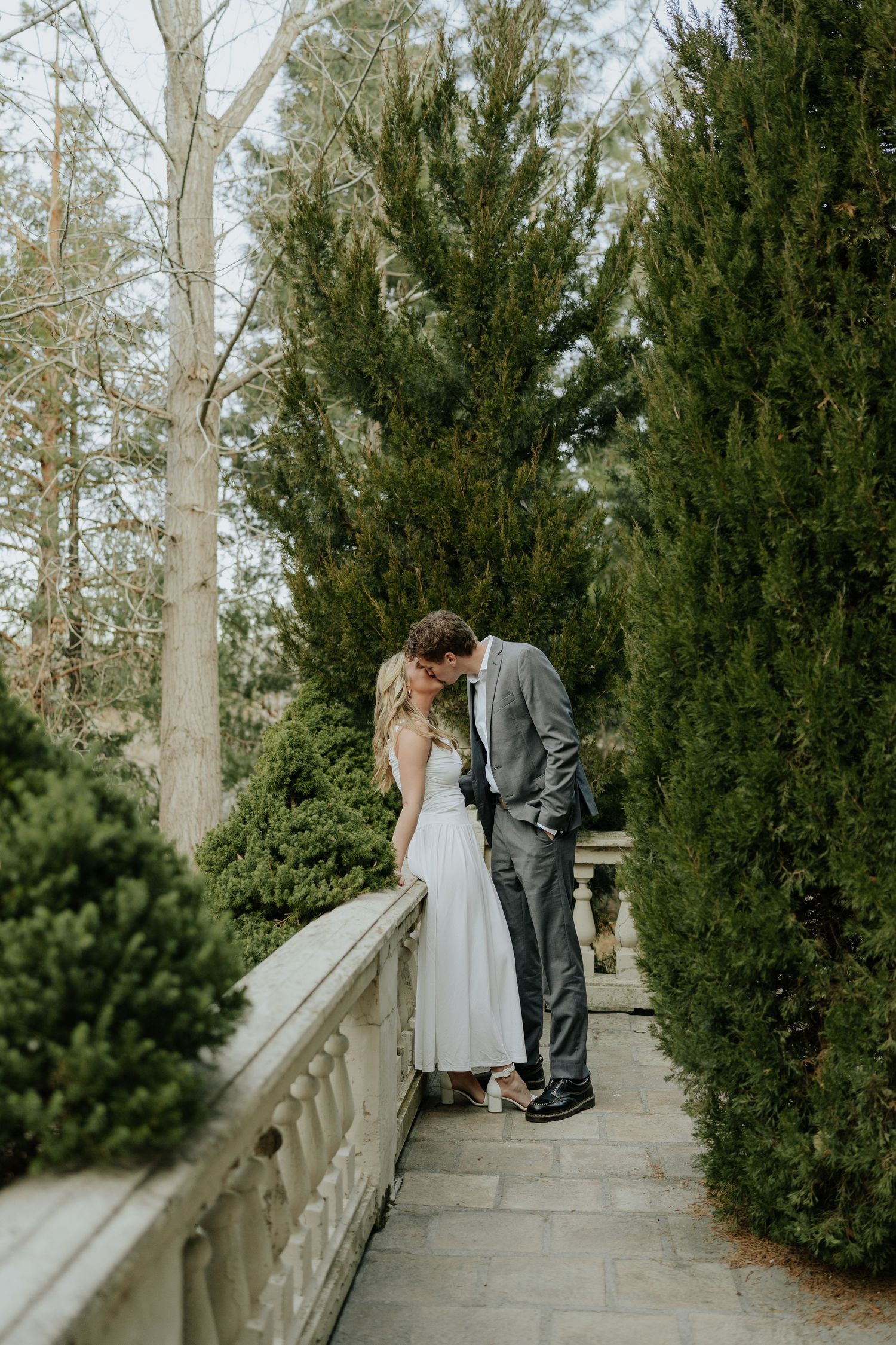 Couple sharing a romantic kiss on a stone pathway lined with tall evergreen trees near a balustrade at La Caille Restaurant in Utah.