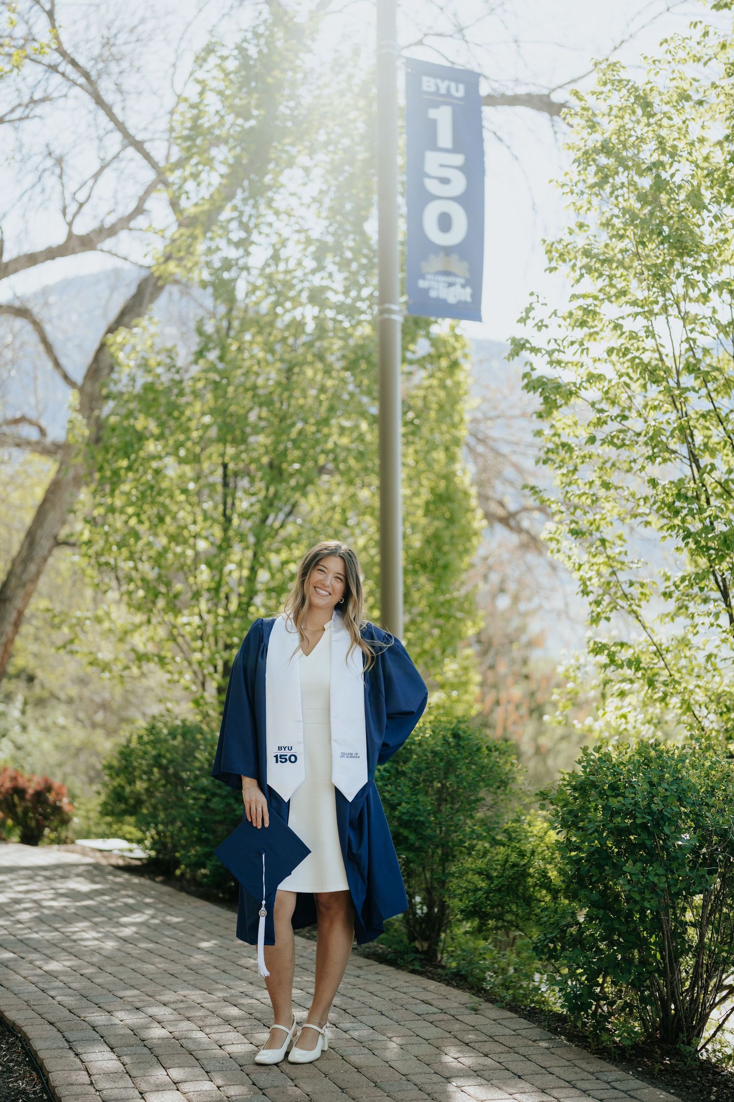 A smiling woman in a navy blue graduation gown and white dress poses on a brick path at BYU's 150th anniversary celebration.