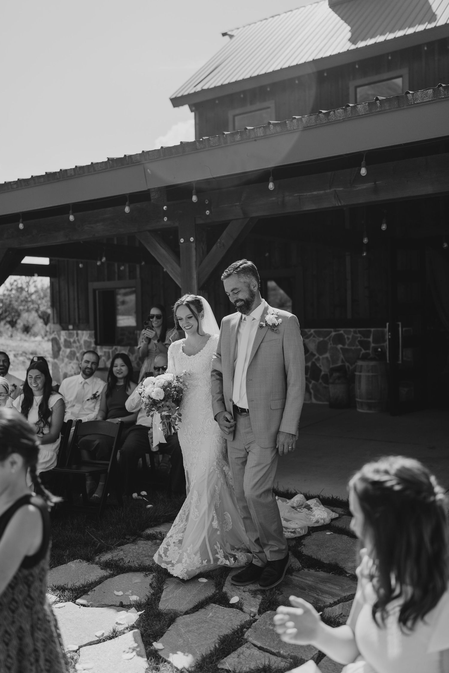 Black and white photo of newlyweds walking down aisle after ceremony at rustic barn venue.
