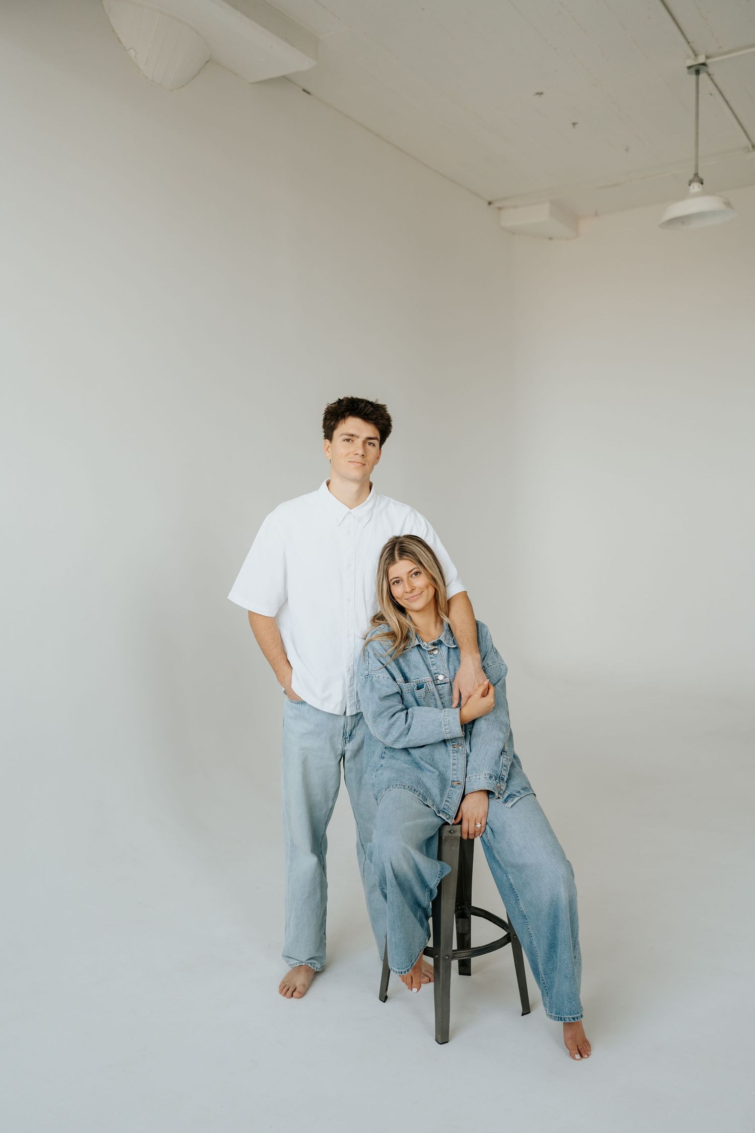 Couple posing at Studio Elevn in Salt Lake City, Utah, woman in denim jumpsuit seated on stool, man standing behind.