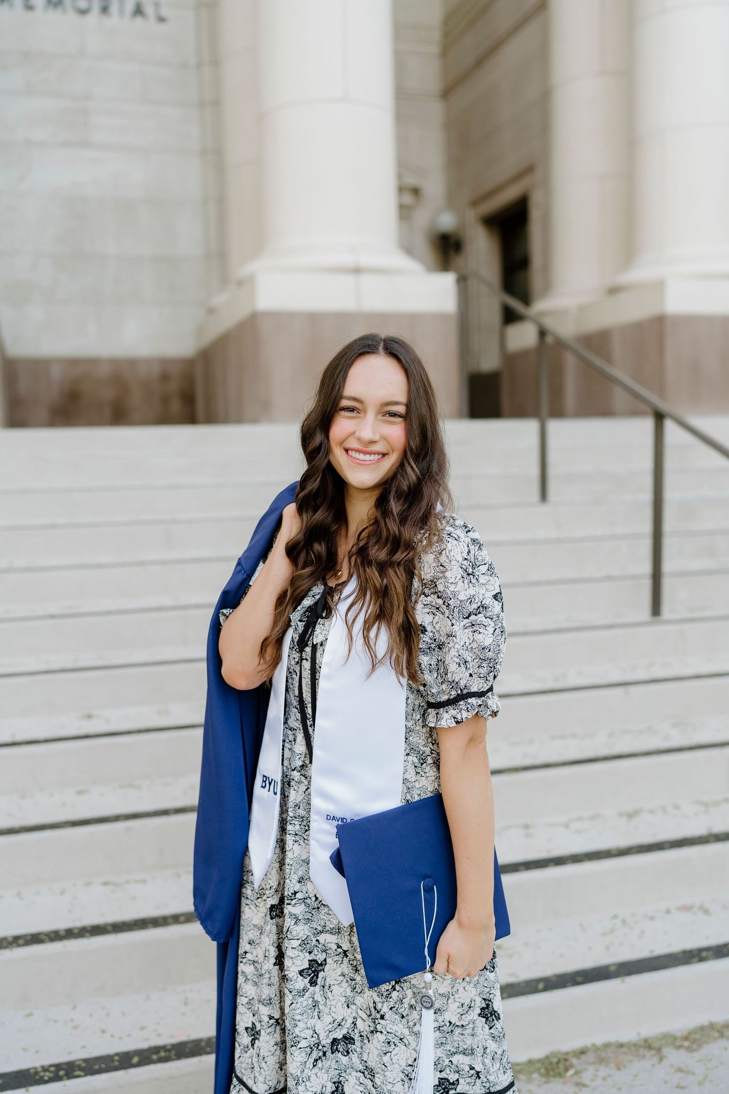 Smiling BYU graduate in floral dress carrying blue gown and diploma on campus steps.