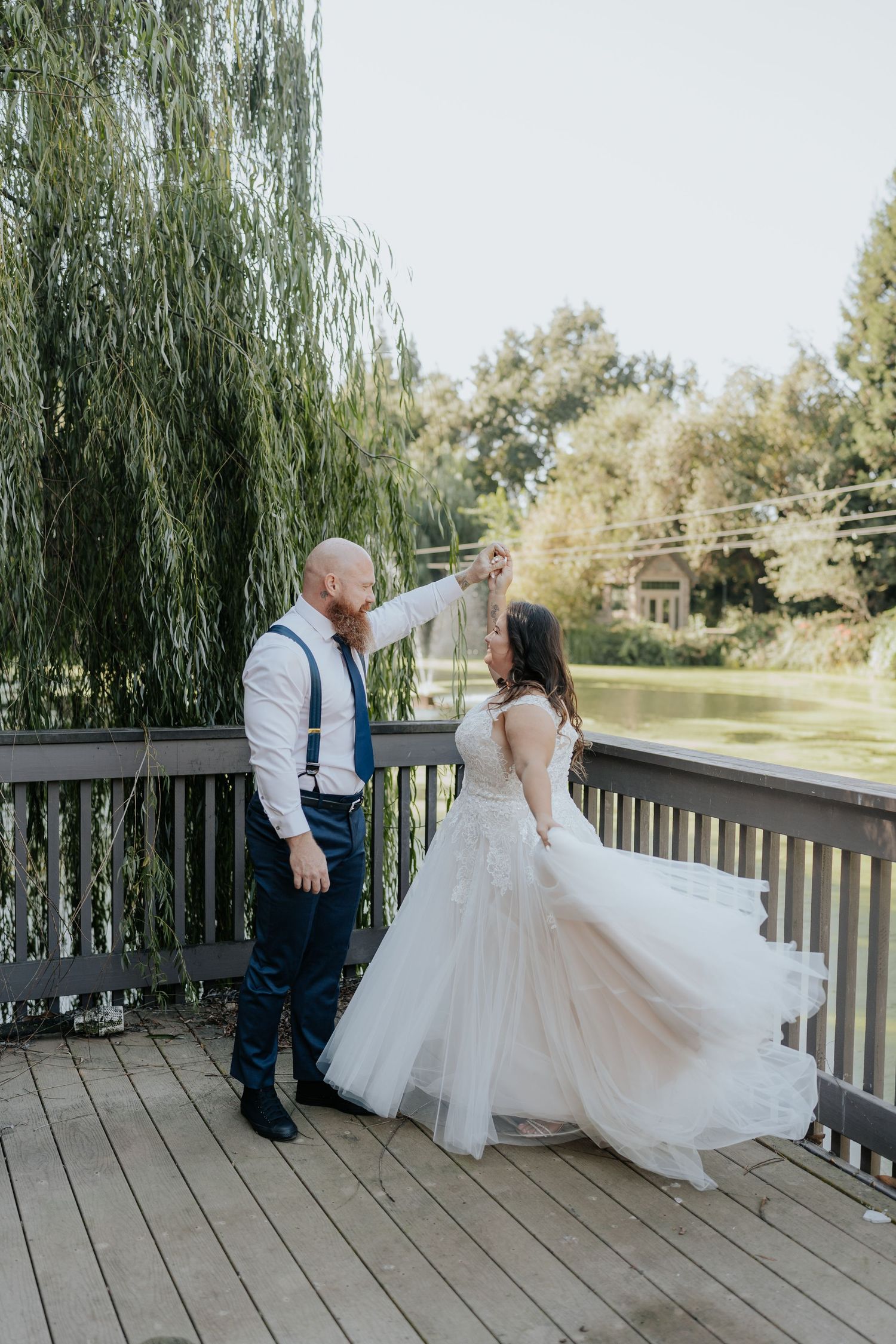 Bride and groom share a romantic dance on a wooden deck overlooking a pond, surrounded by weeping willow trees.