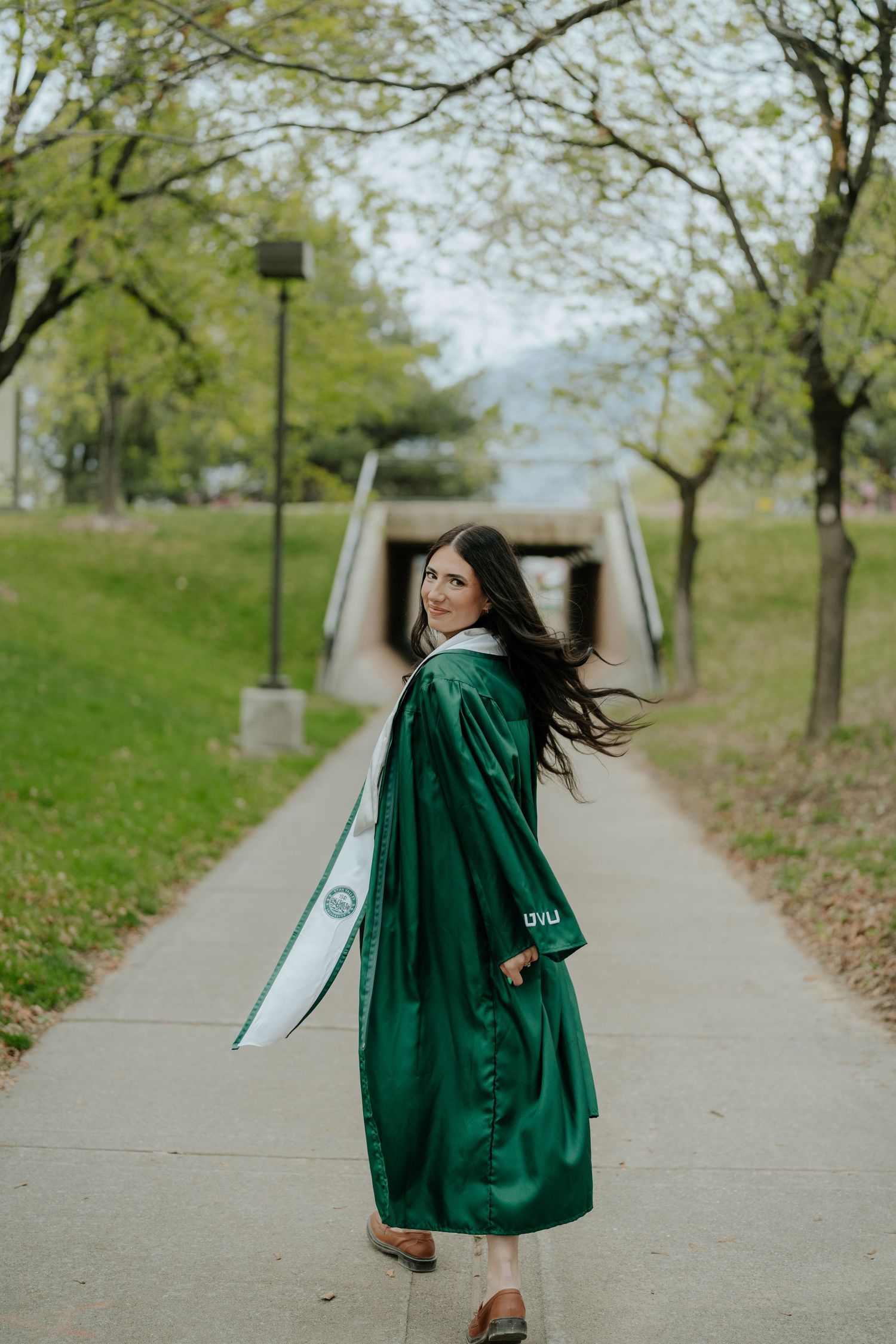 A woman in a green graduation gown and sash walks along a tree-lined campus path, glancing back over her shoulder.