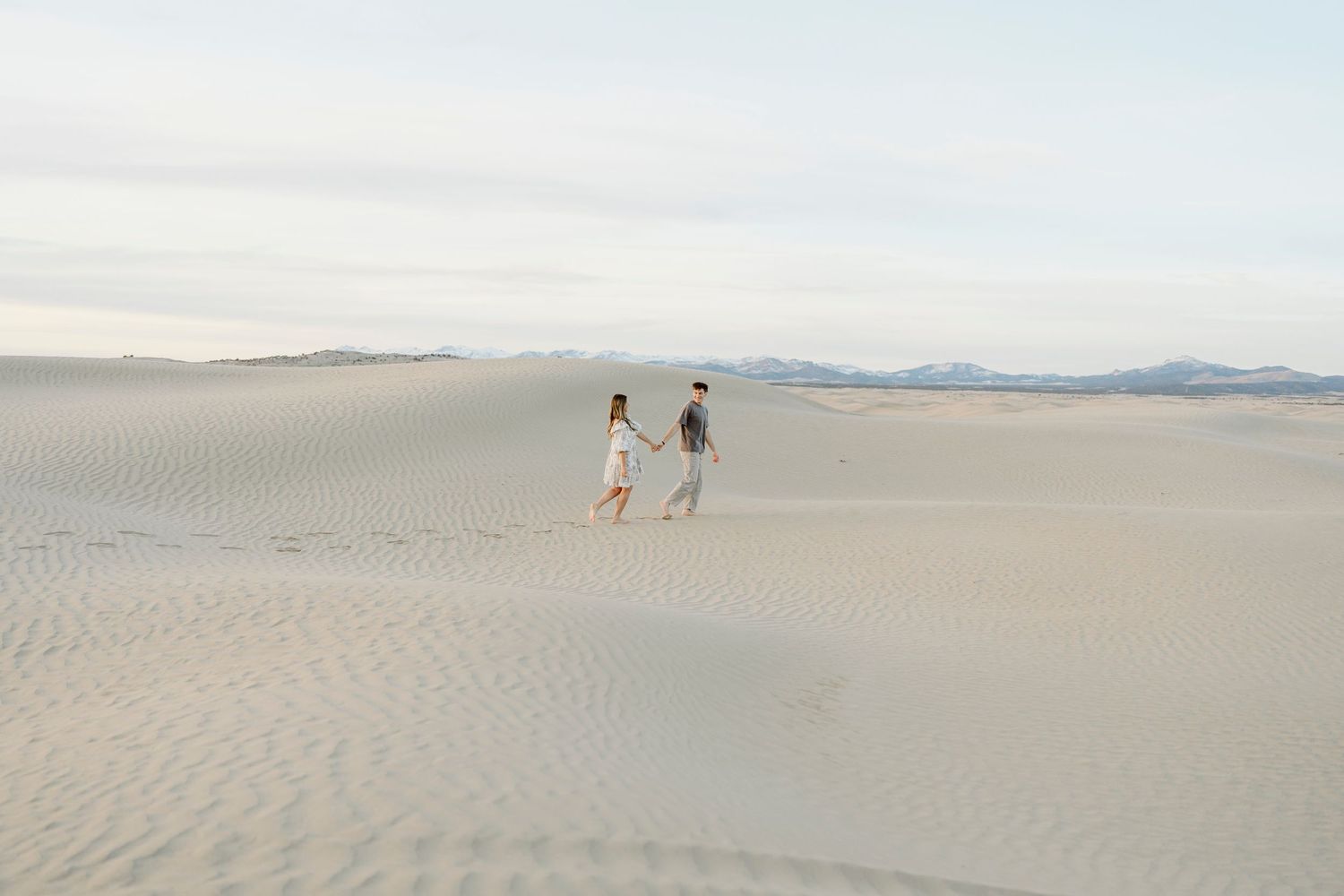 Couple holding hands while walking across vast white sand dunes under a soft sky.