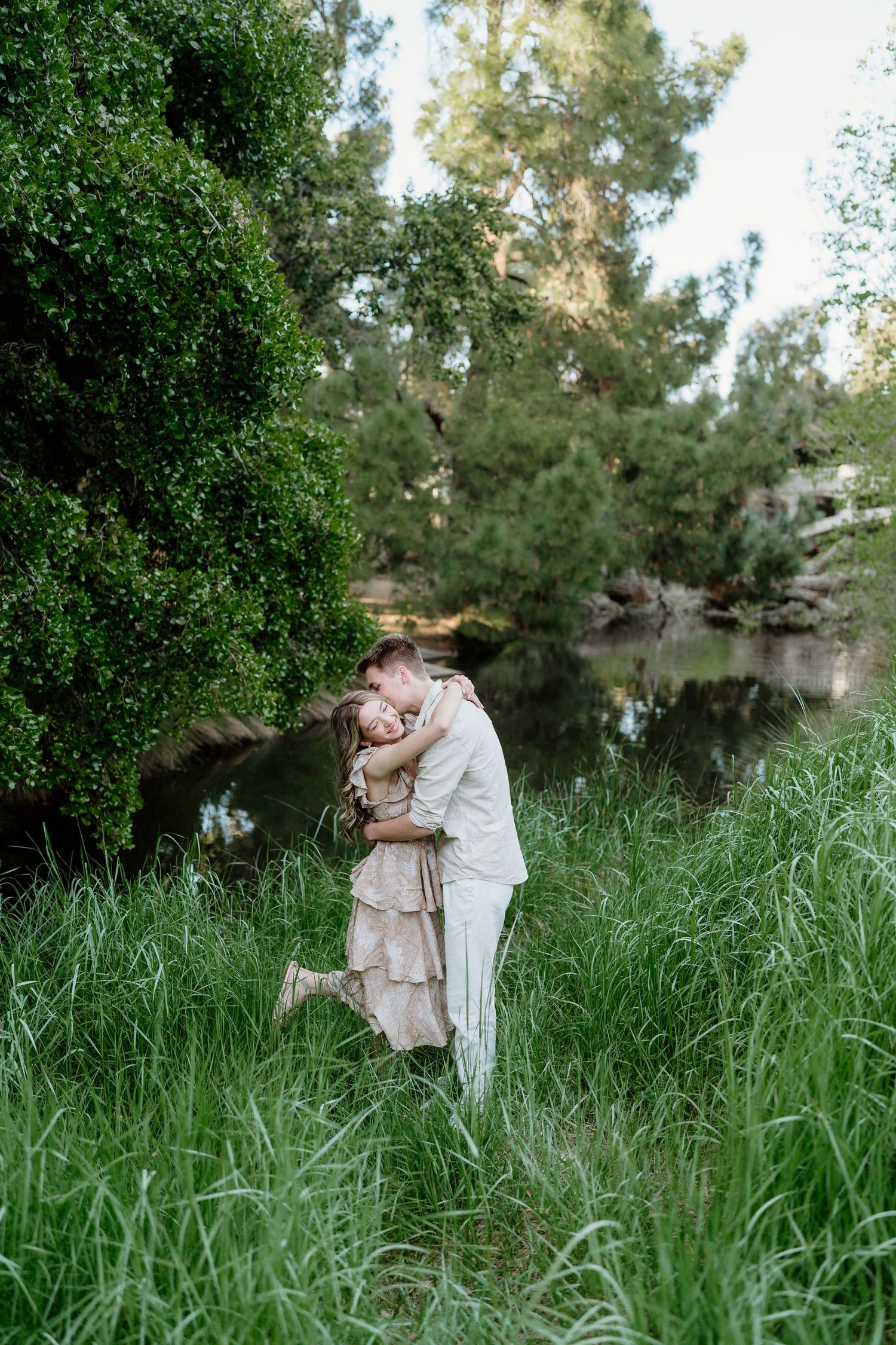 Couple sharing a romantic embrace in lush green grass beside a tranquil woodland pond.
