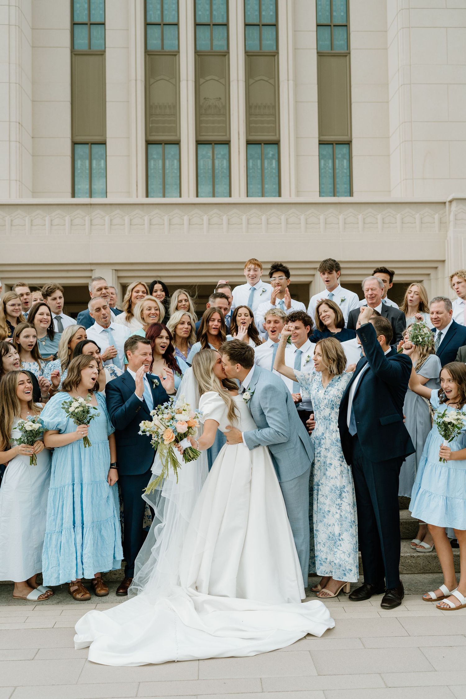 Bride and groom share a kiss surrounded by cheering wedding guests on the steps of a temple.