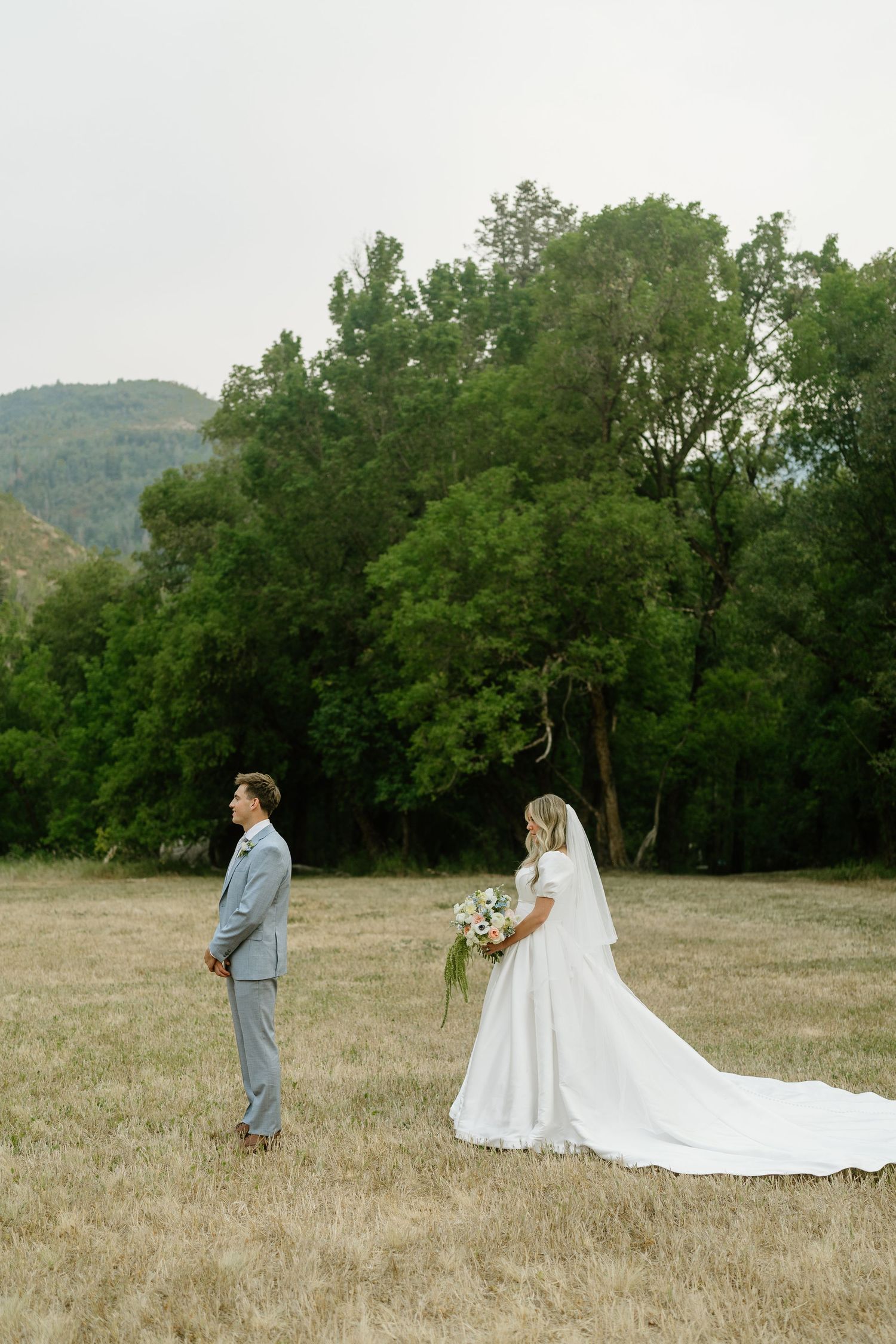 Bride in white gown with long train and groom in gray suit stand in golden field with green trees and mountains.