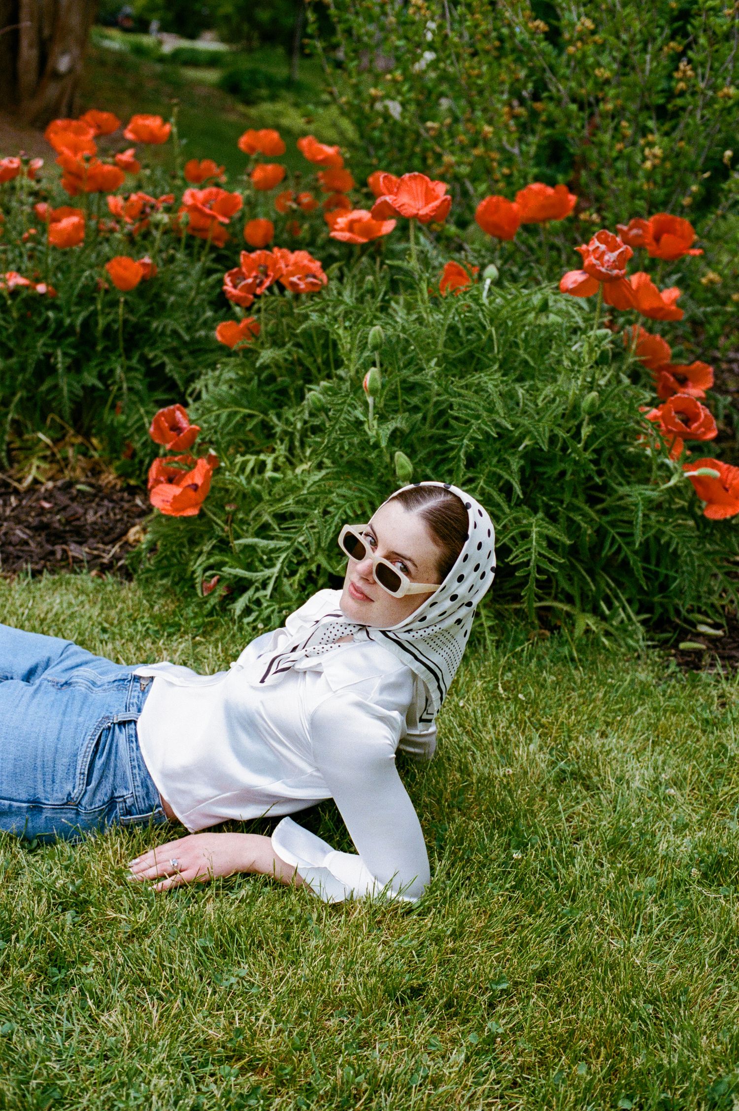 Woman in white top, jeans, polka dot headscarf and sunglasses lying on grass beside vibrant red poppy flowers.
