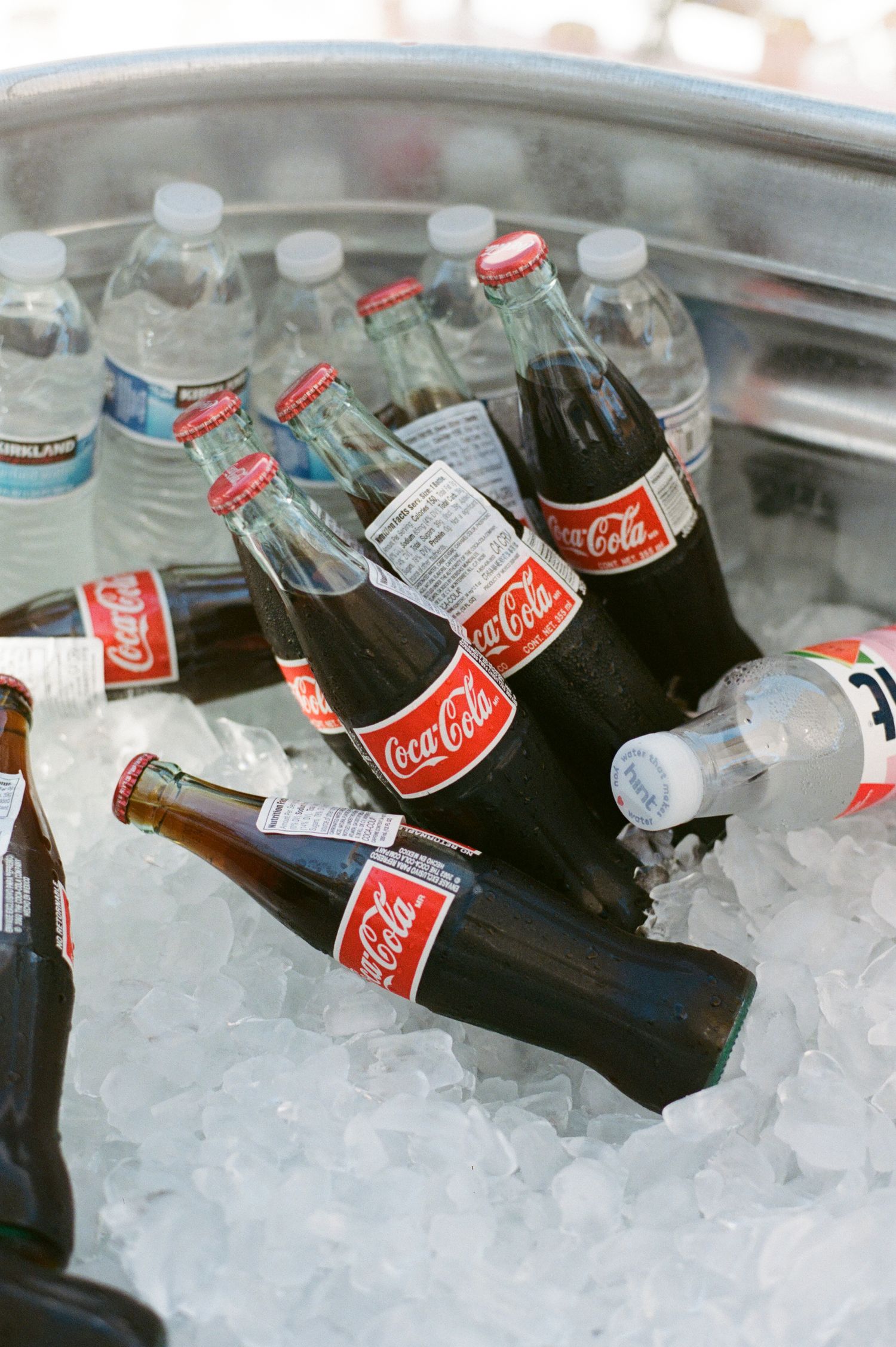 Glass Coca-Cola bottles and water bottles chilling on ice in a large metal cooler at an outdoor event.