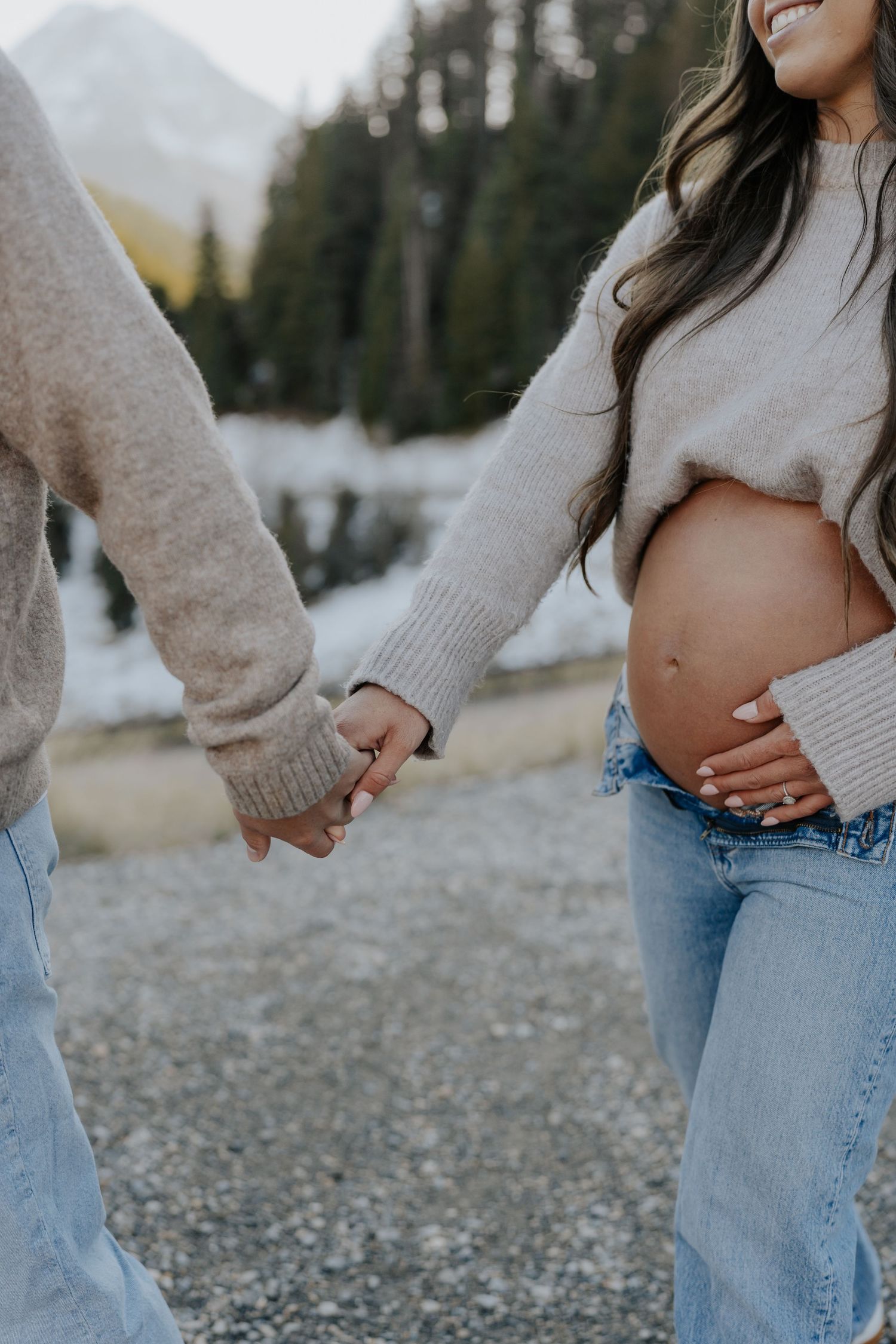Pregnant couple holding hands outdoors in winter, woman cradling baby bump in cropped sweater and jeans.