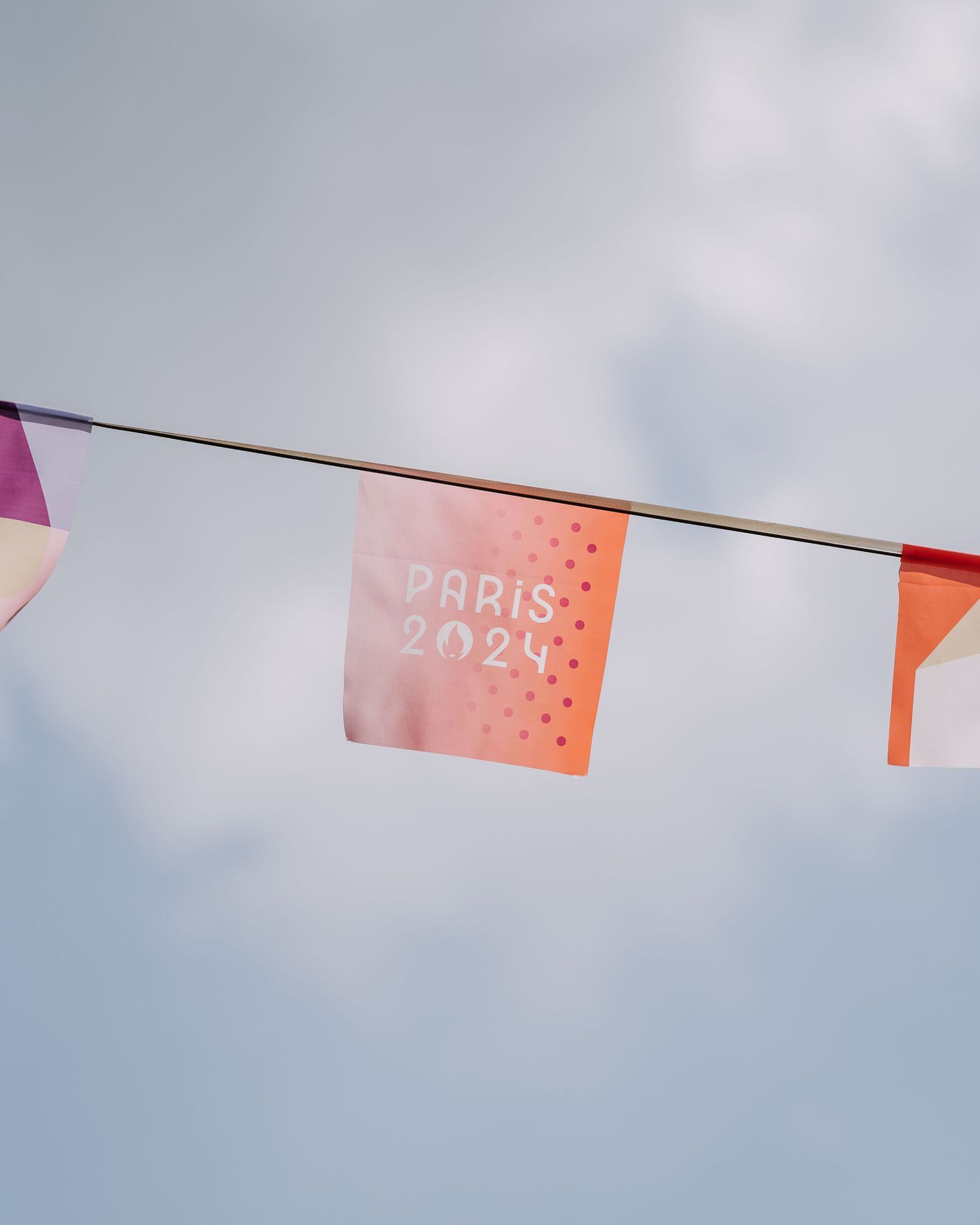 Colorful Paris 2024 Olympics bunting flags hanging against a cloudy blue sky.