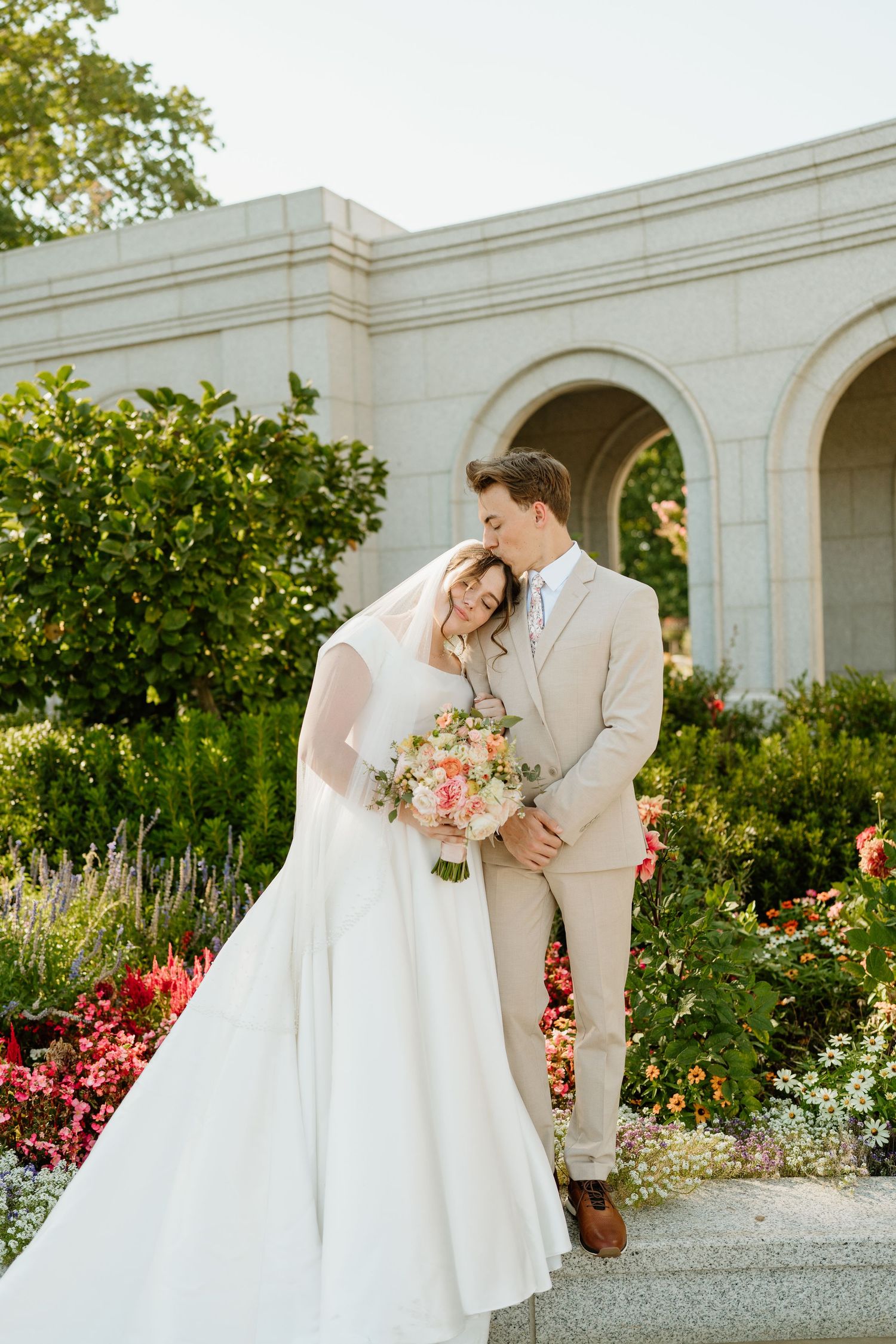 Groom kissing bride on head in lush garden setting outside elegant white stone building with arches.