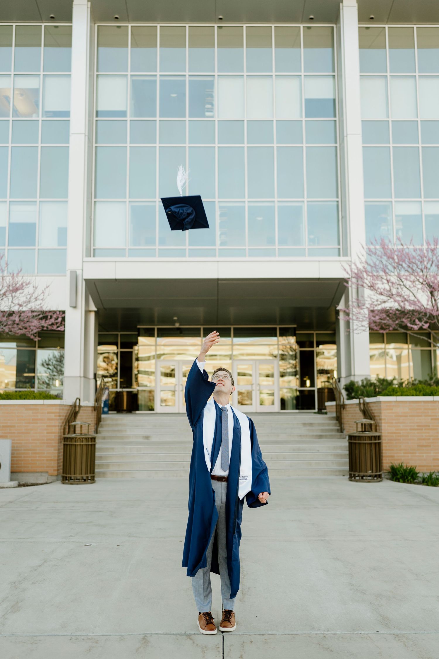 BYU graduate in blue gown tossing mortarboard cap in front of campus building.