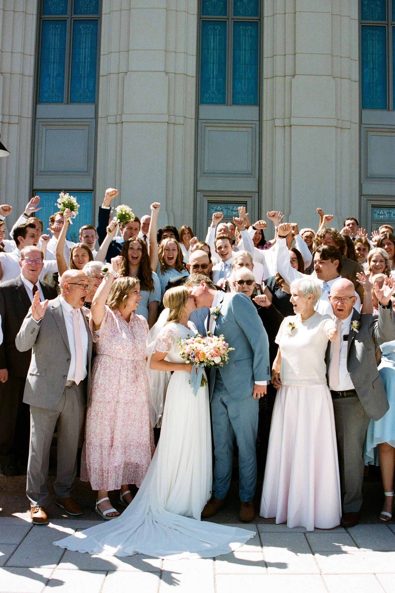 A newlywed couple shares a kiss surrounded by cheering guests on church steps in bright sunlight.