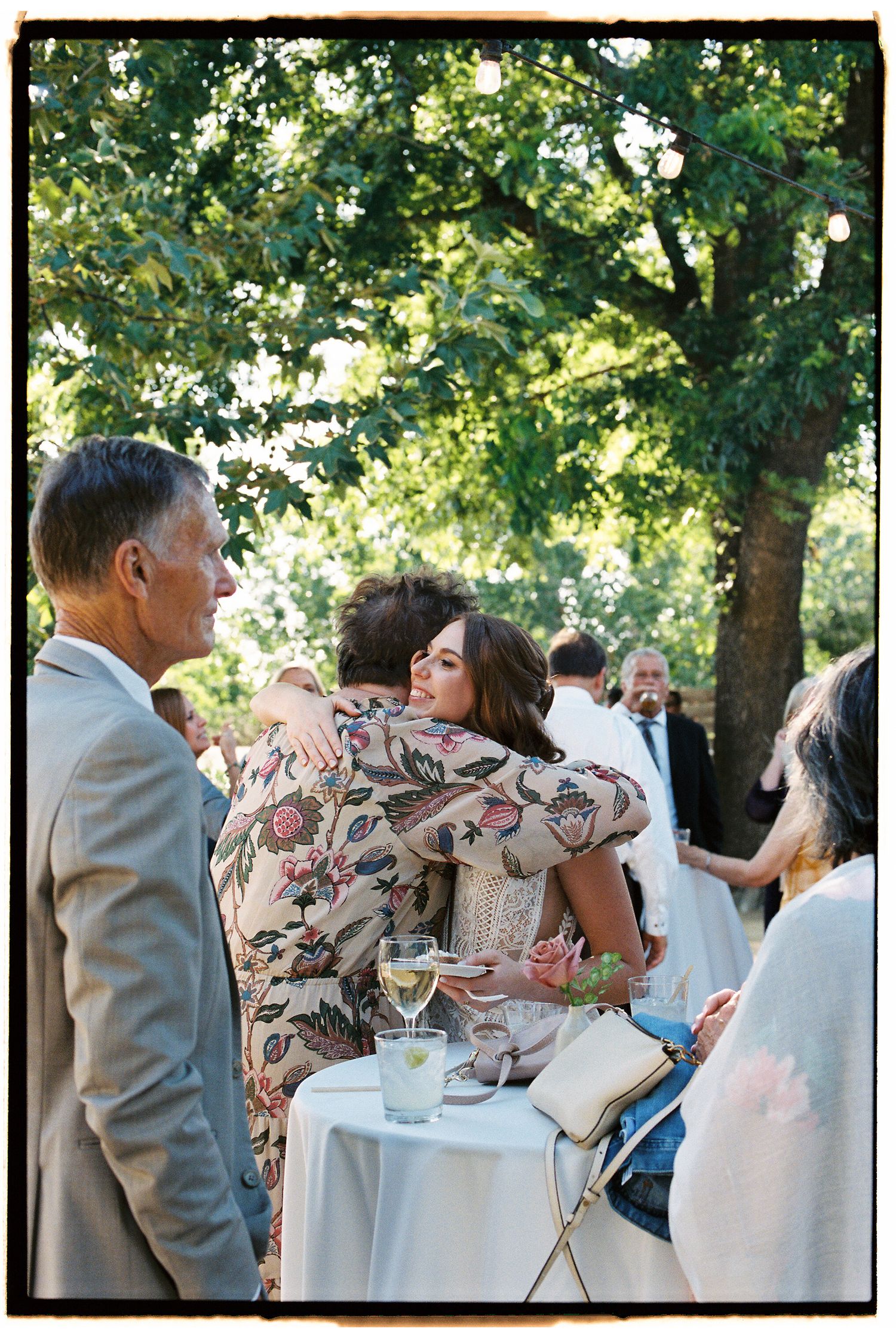 Wedding guests embracing and socializing outdoors under lush green trees at a garden reception.