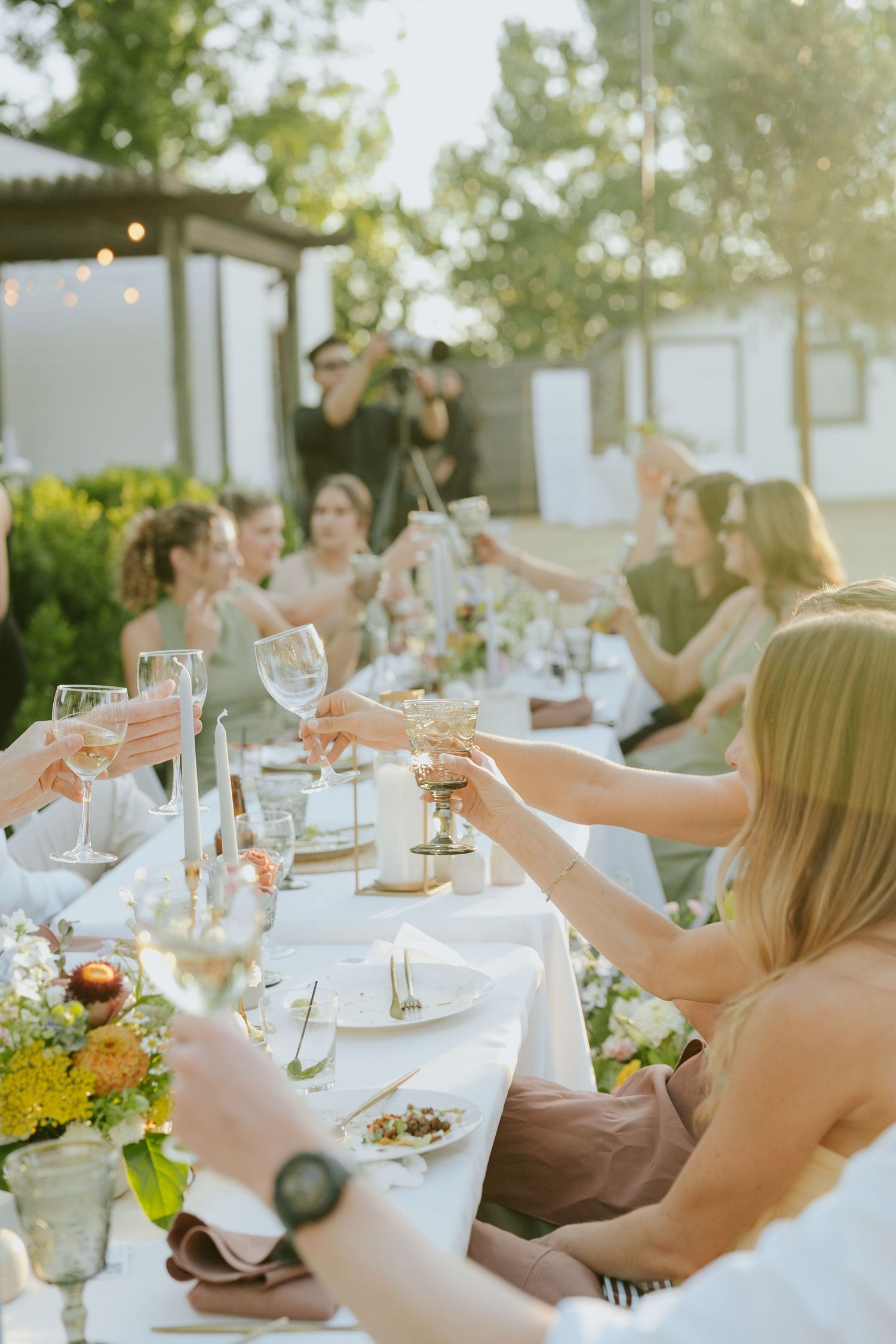 Guests raising wine glasses in a toast at an elegant outdoor wedding reception dinner table with floral arrangements.