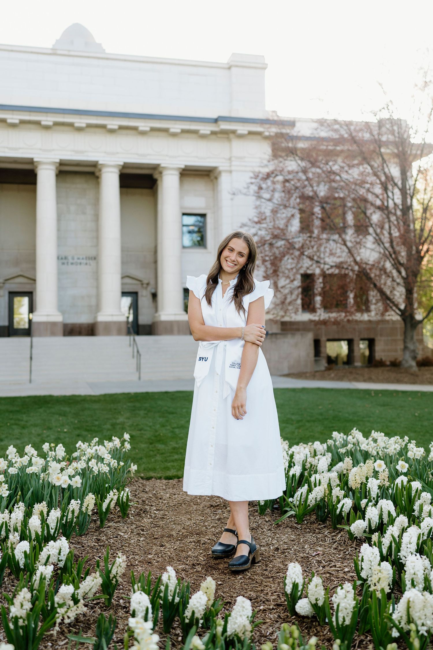Young woman in white dress standing among white flowers outside BYU Maeser Building.