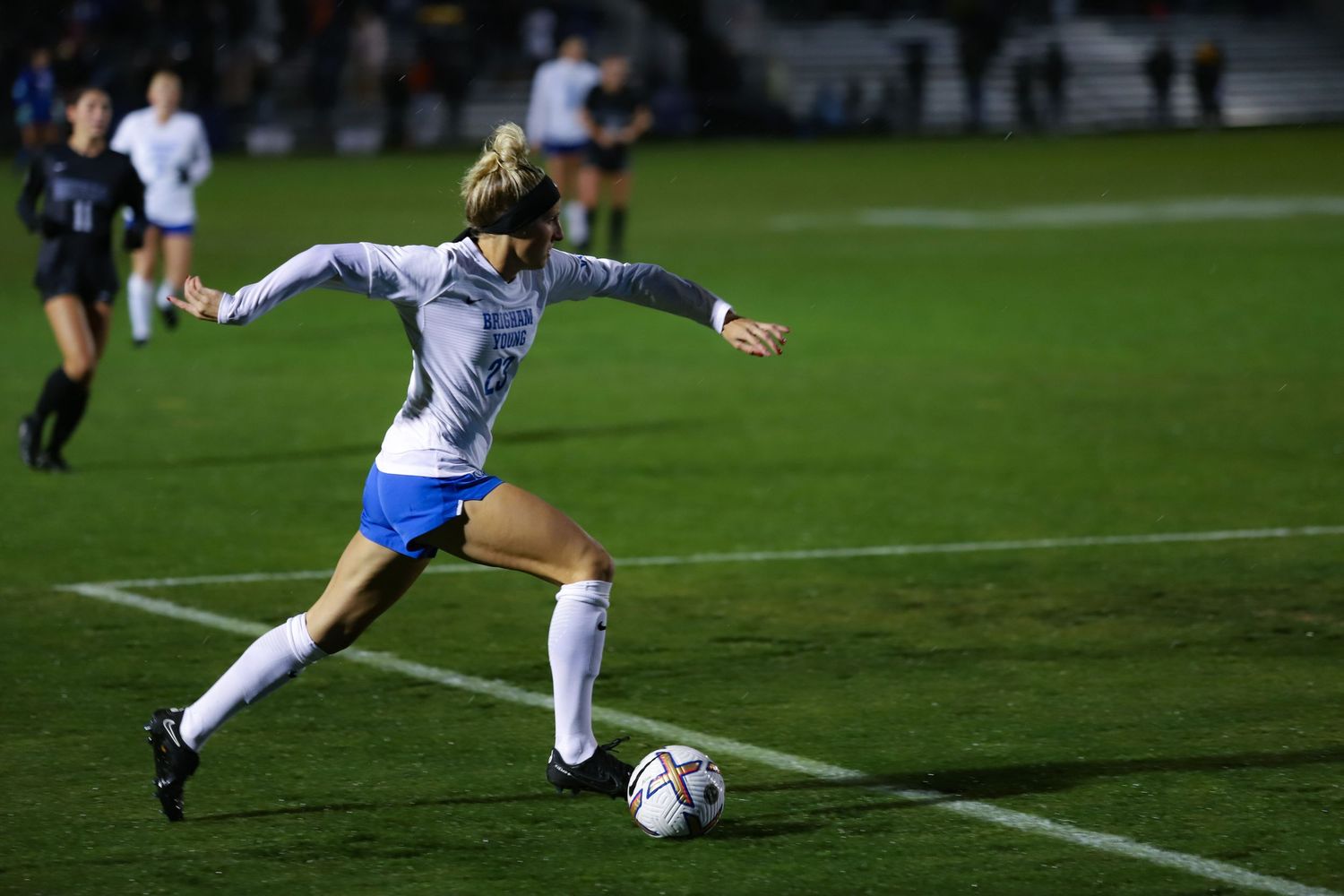 A female soccer player in white and blue uniform prepares to kick a ball during a night game.