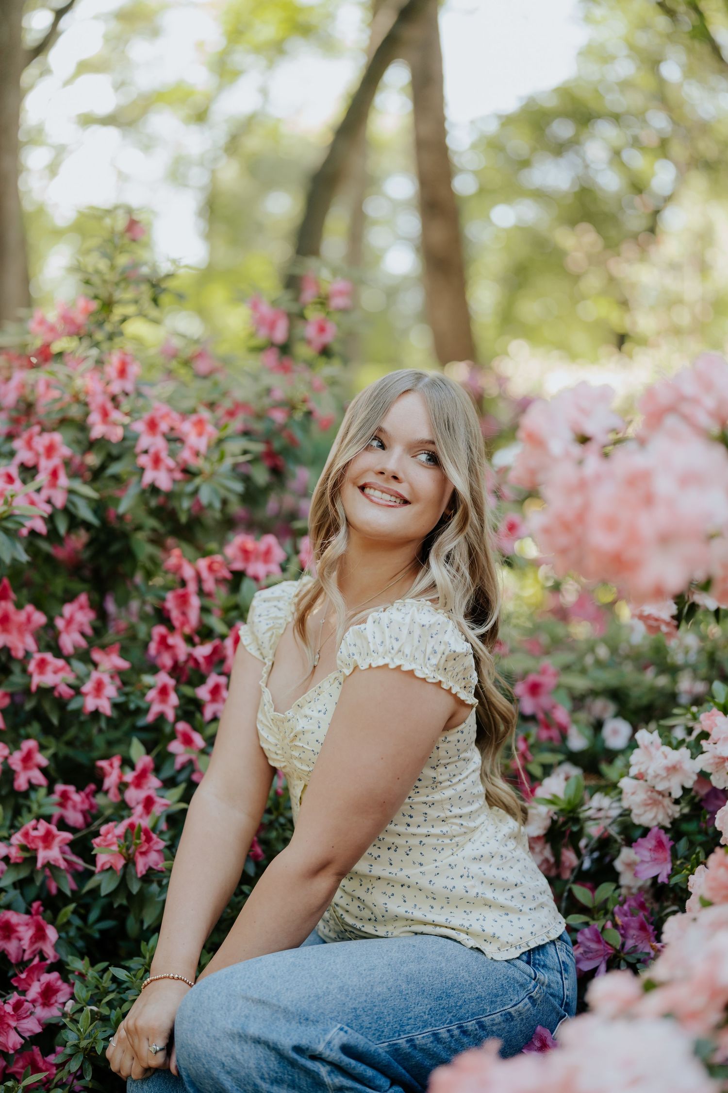 Happy blonde girl in floral top sitting among vibrant pink azalea blooms in garden near Sacramento, California.