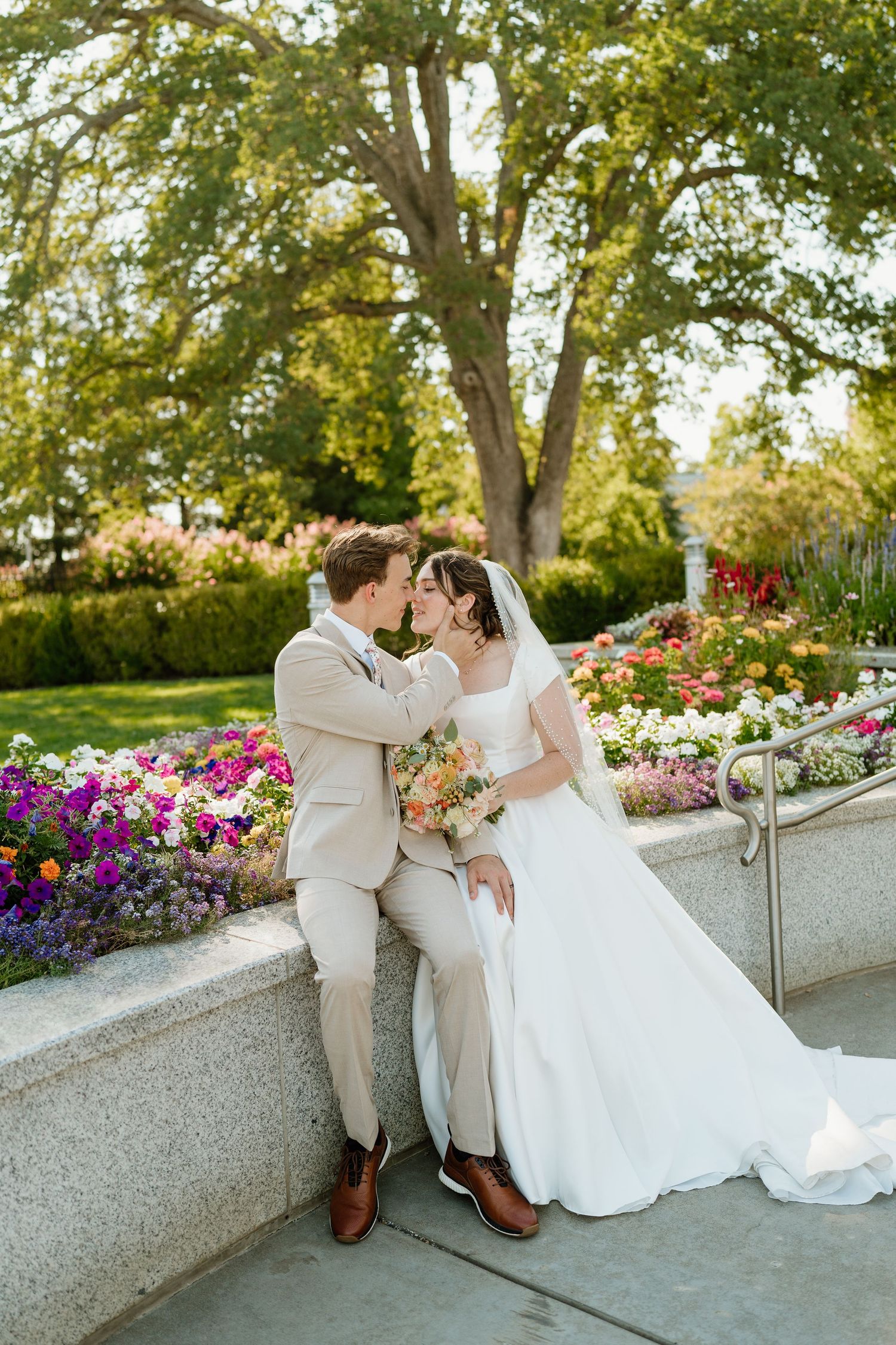 Bride and groom share a kiss beside colorful garden flowers, groom in tan suit with bouquet at Sacramento LDS temple.