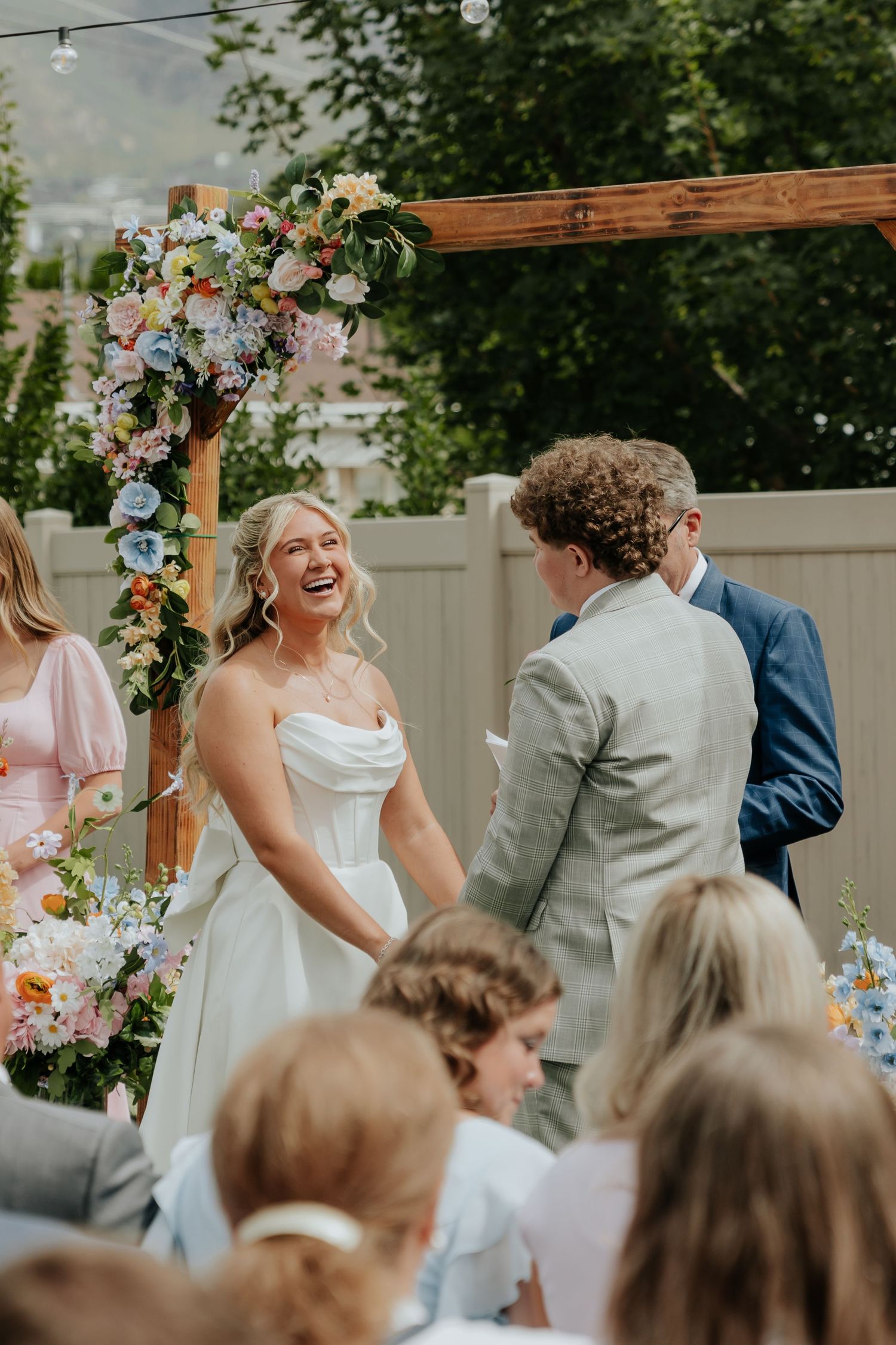 Radiant bride laughs joyfully at outdoor ceremony beneath a colorful floral wooden arch.