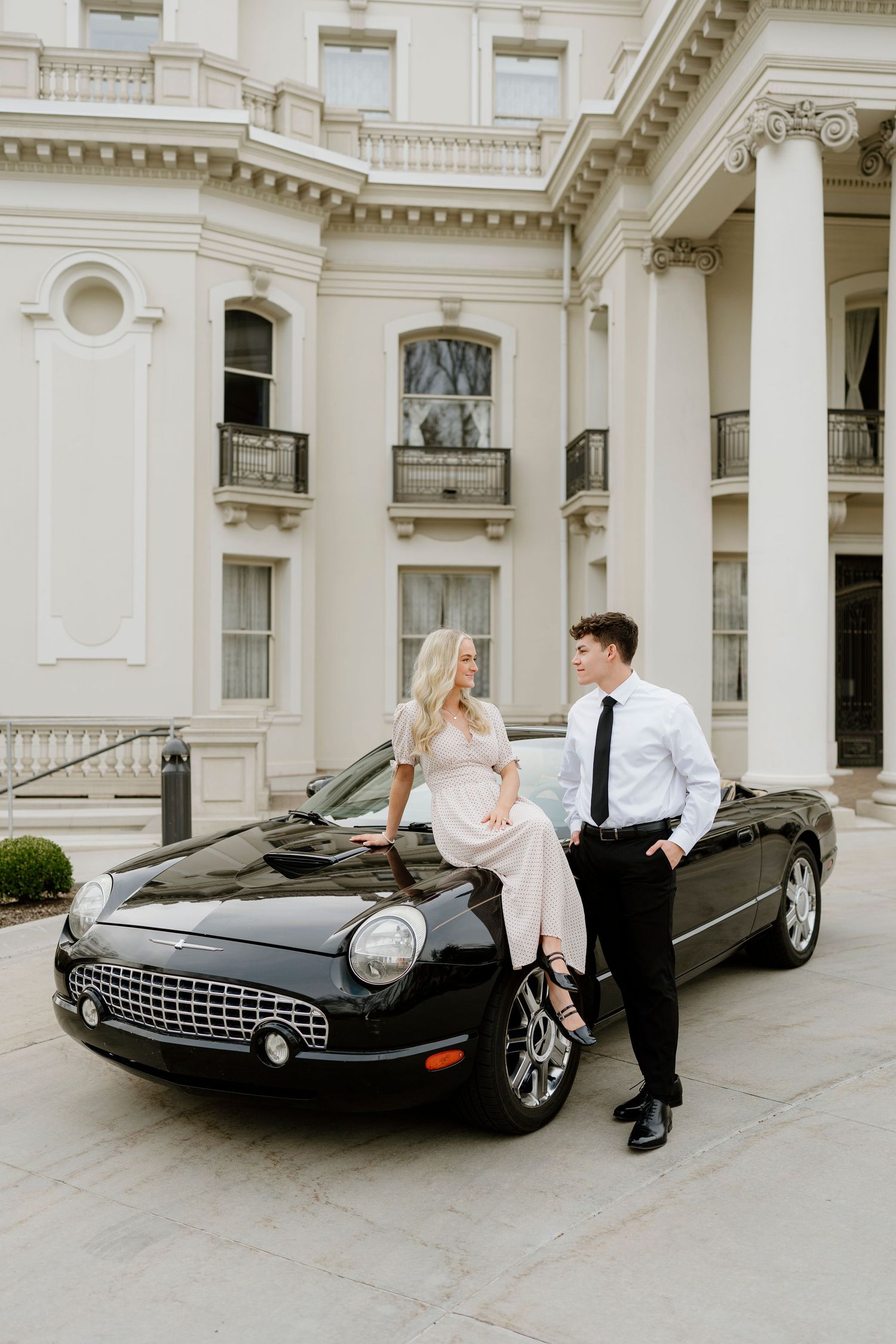 Elegant couple posing beside a black convertible Thunderbird in front of a grand white mansion.