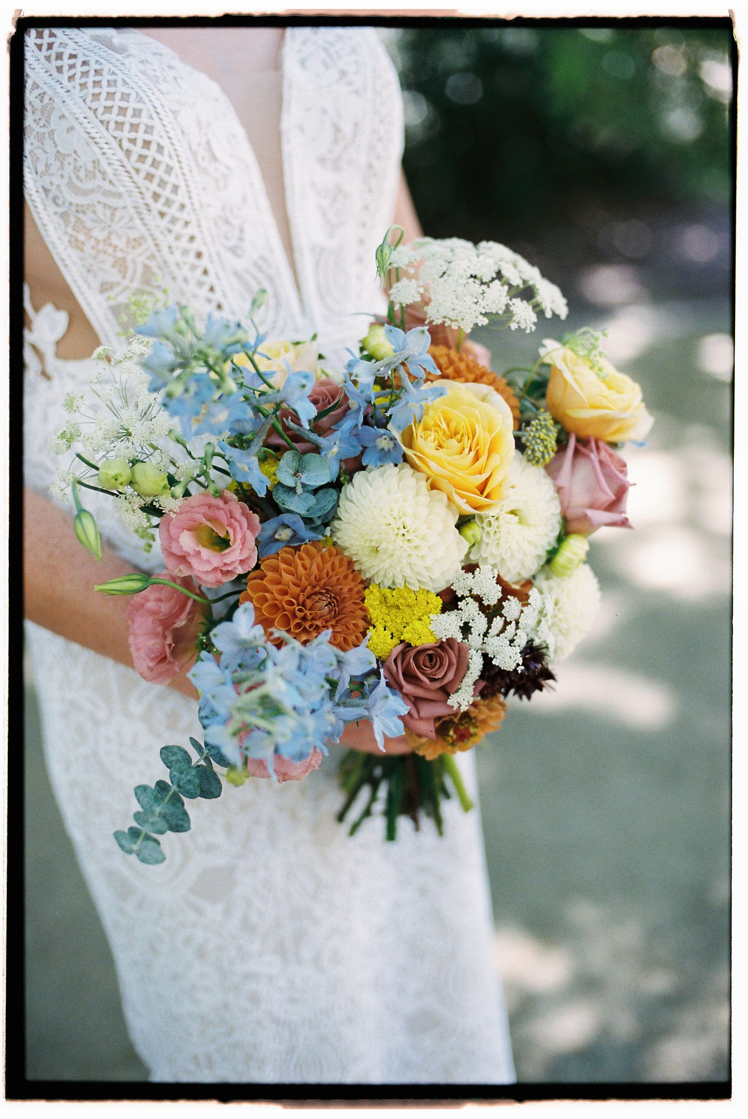 Bride holding a colorful wedding bouquet with yellow roses, blue hydrangeas, pink ranunculus, and orange dahlias.