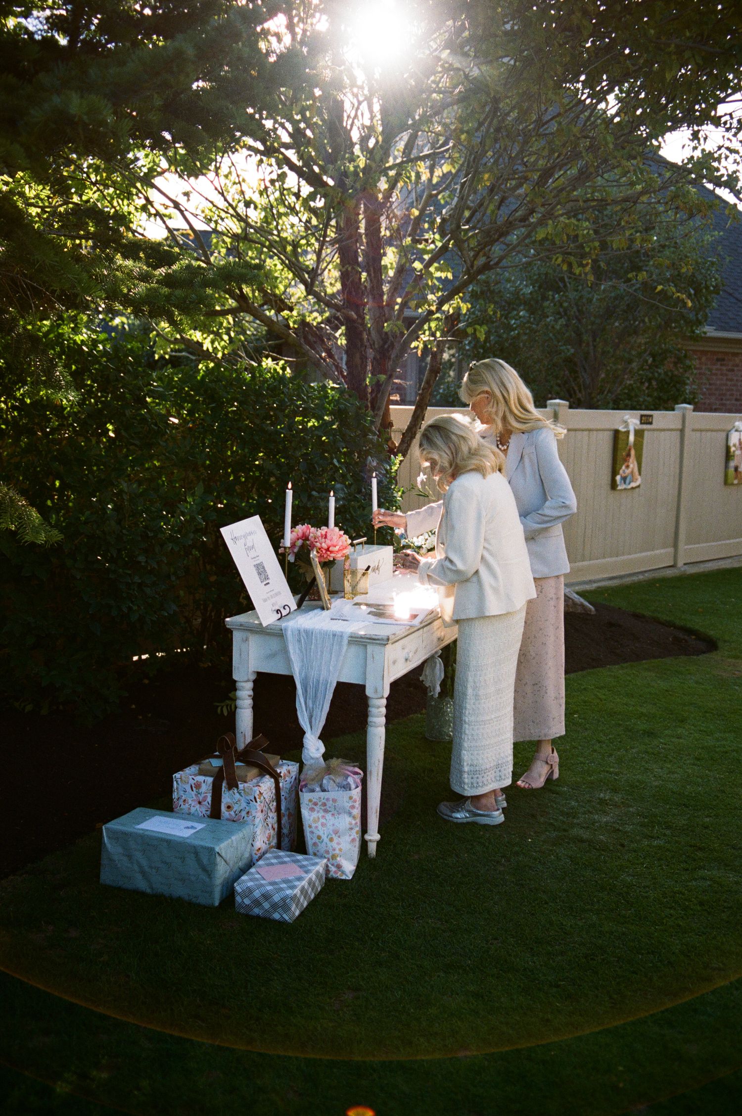 Two women stand at an elegant white outdoor table with gifts and floral arrangements at a garden party.