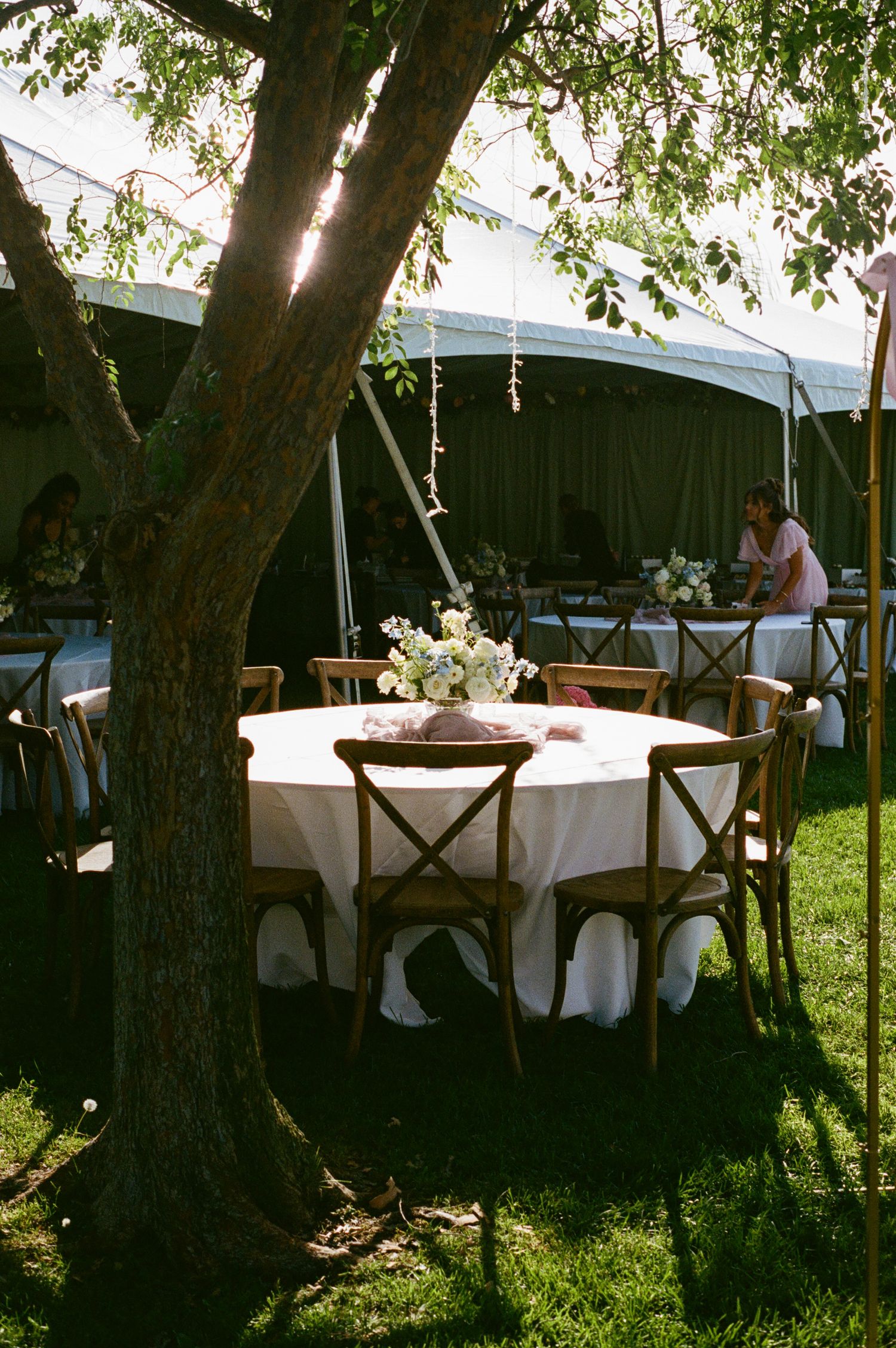 Elegant outdoor wedding reception with round tables, wooden cross-back chairs, and floral centerpieces under a white tent.